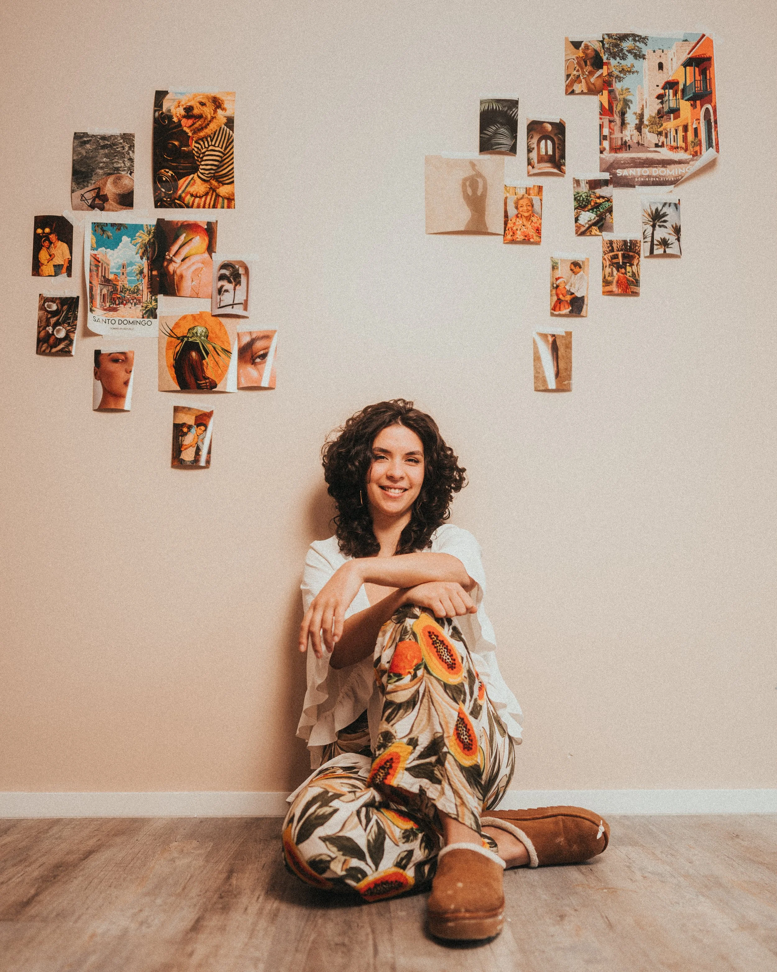 A woman with curly hair smiling and sitting on the floor with her legs crossed, leaning against a wall decorated with various colorful photos and posters. She is wearing a white top and floral pants, with brown boots, and is sitting on a wooden floor.
