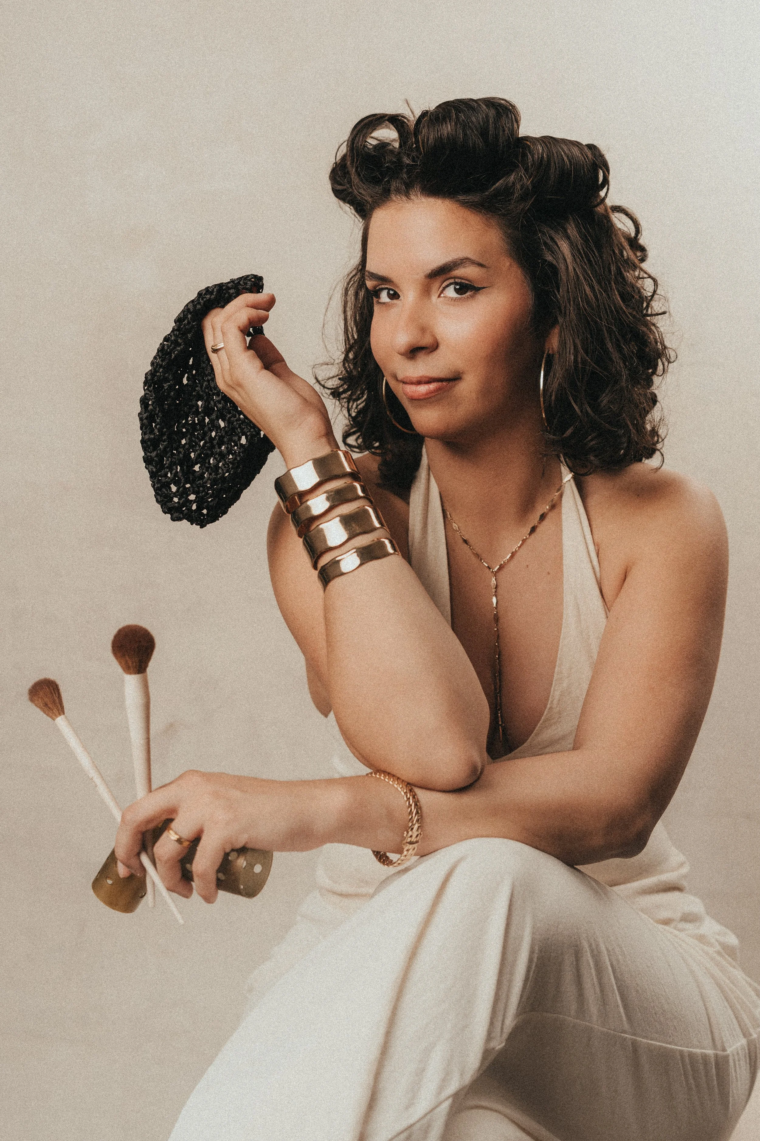 Young woman with curly hair styled in vintage rolls, wearing hoop earrings, layered bracelets, and a beige dress, sitting on a light-colored sofa, holding a makeup brush and a glittery black makeup bag, with a neutral background.