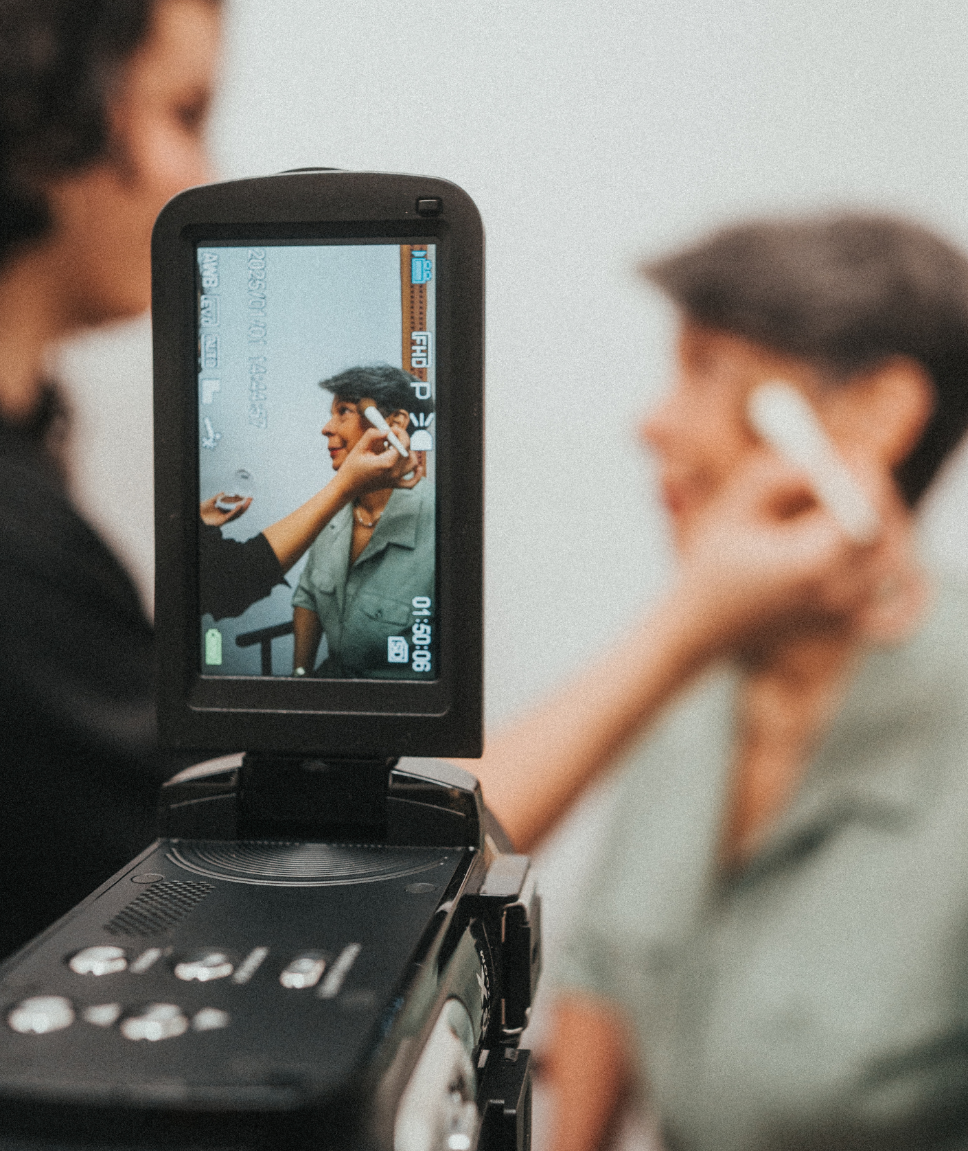 A person is using a camera on a tripod to take a photo of a woman with short dark hair, wearing a green shirt, in front of a whiteboard with text and writing on it.