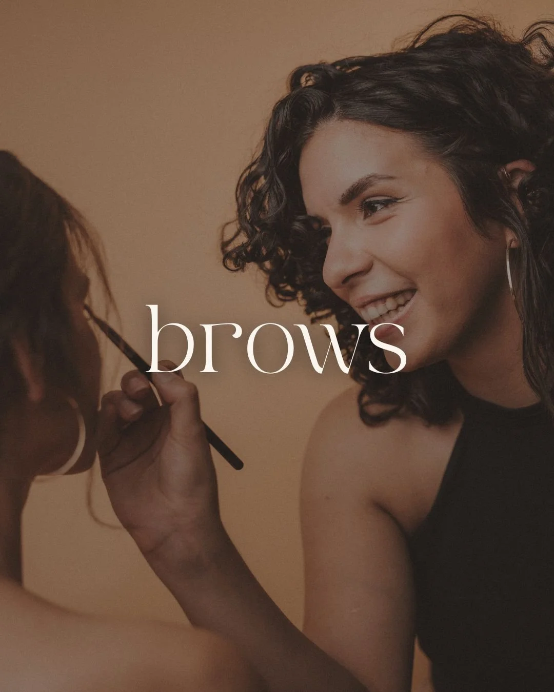 A woman with curly hair smiling as makeup artist applies eyebrow makeup, with the word 'brows' overlayed in the center.