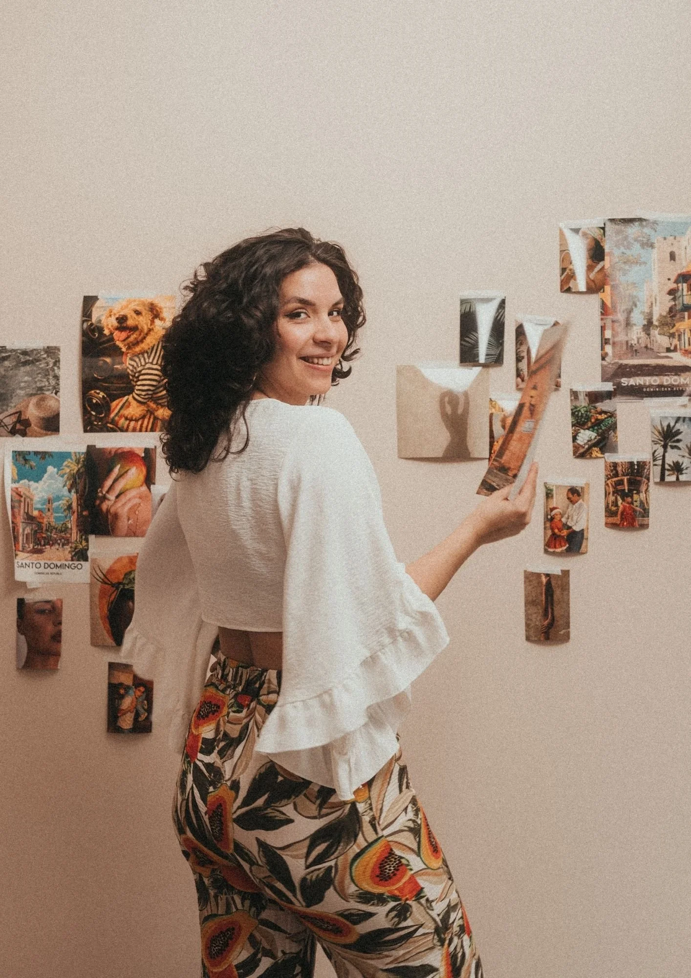 A woman with curly dark hair smiling and looking over her shoulder, standing in front of a wall decorated with photos and artwork, wearing a white top and colorful patterned pants.