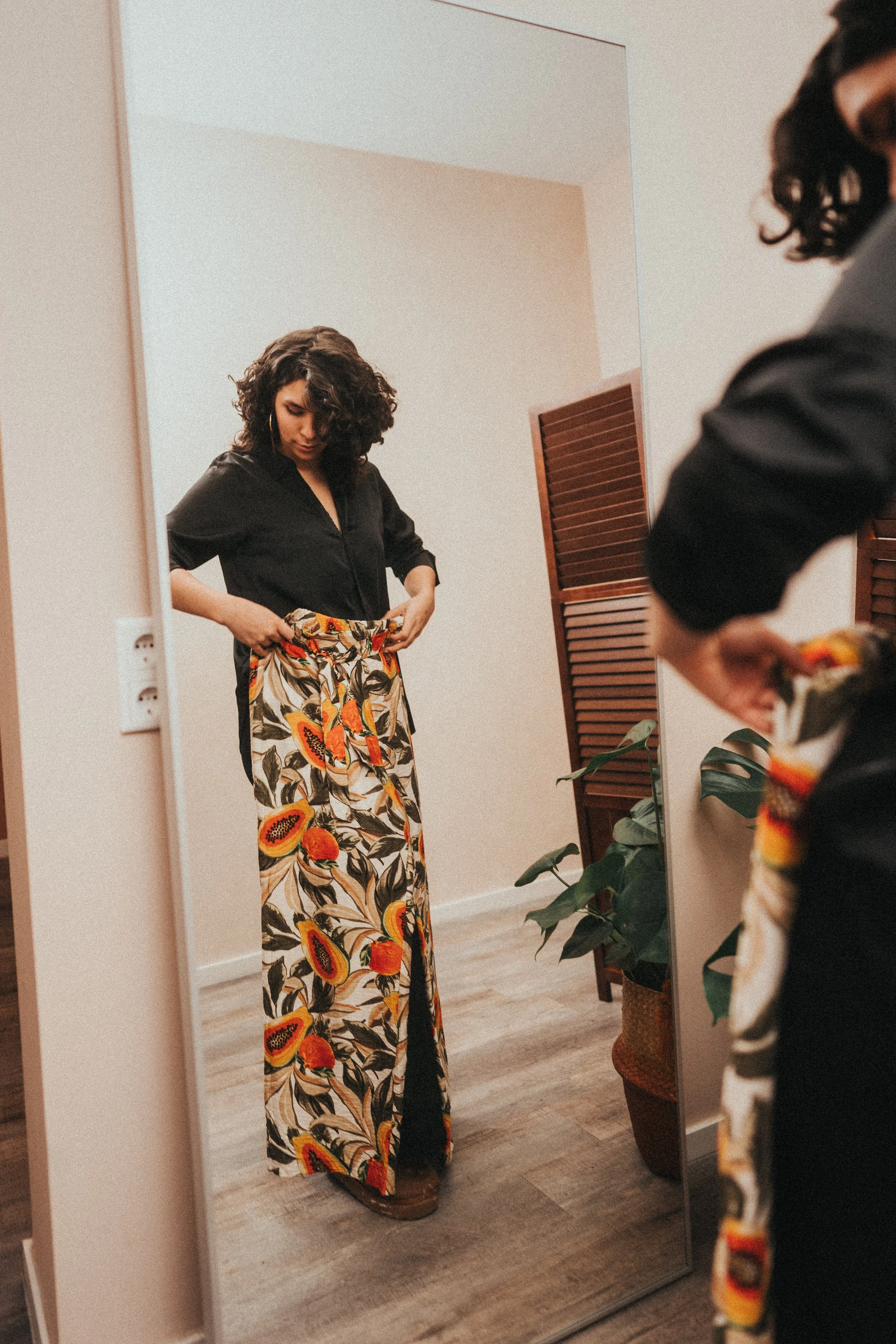 A woman standing in front of a mirror, adjusting her tropical print pants. She has curly dark hair and is wearing a black top. The room has a wooden floor, a plant, and a wooden folding screen in the background.