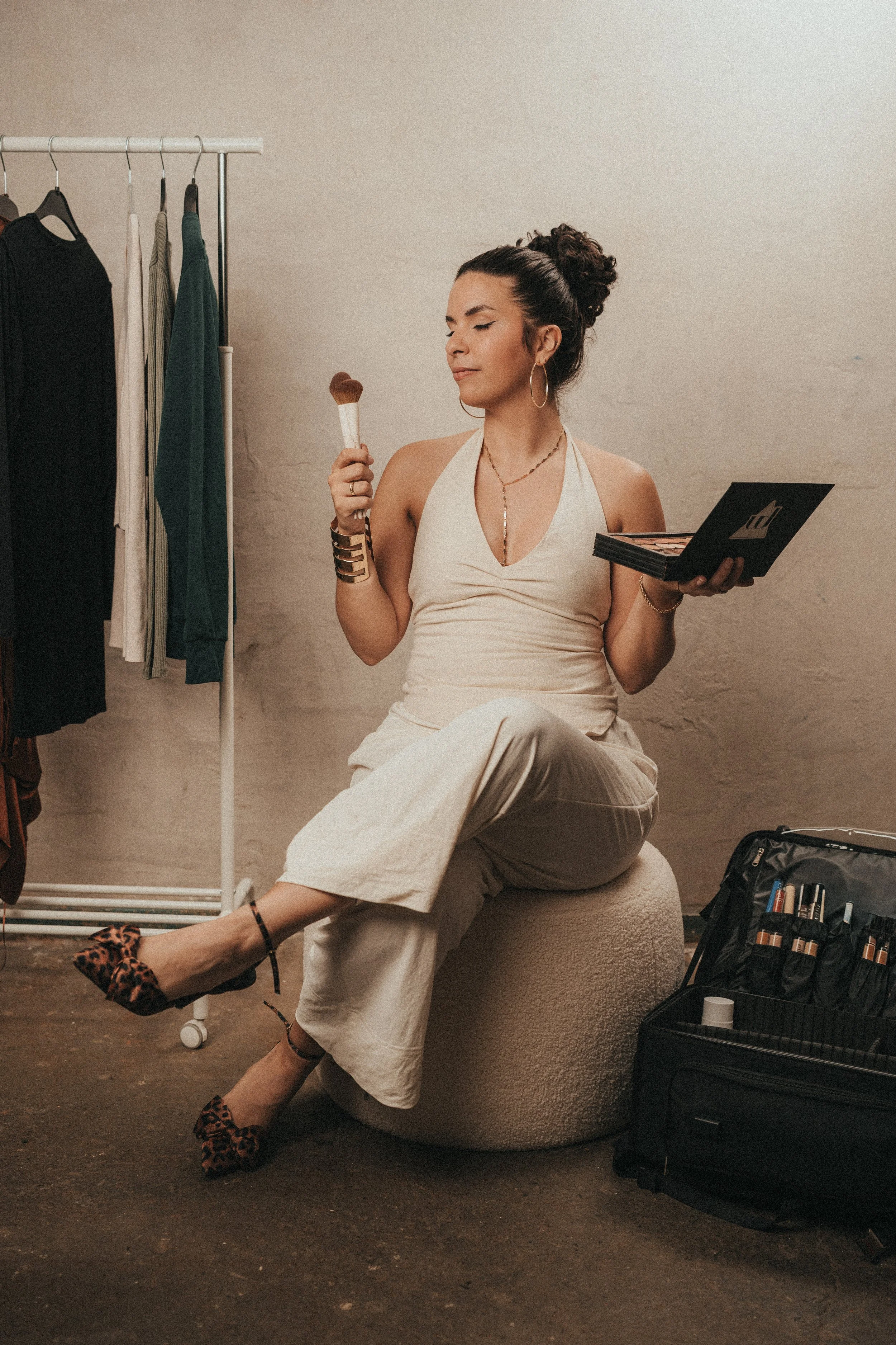 Woman sitting on a round ottoman, holding makeup brushes in one hand and a makeup palette in the other, wearing a sleeveless white top and wide-legged pants, leopard print high heels, in a room with clothing rack and makeup supplies.