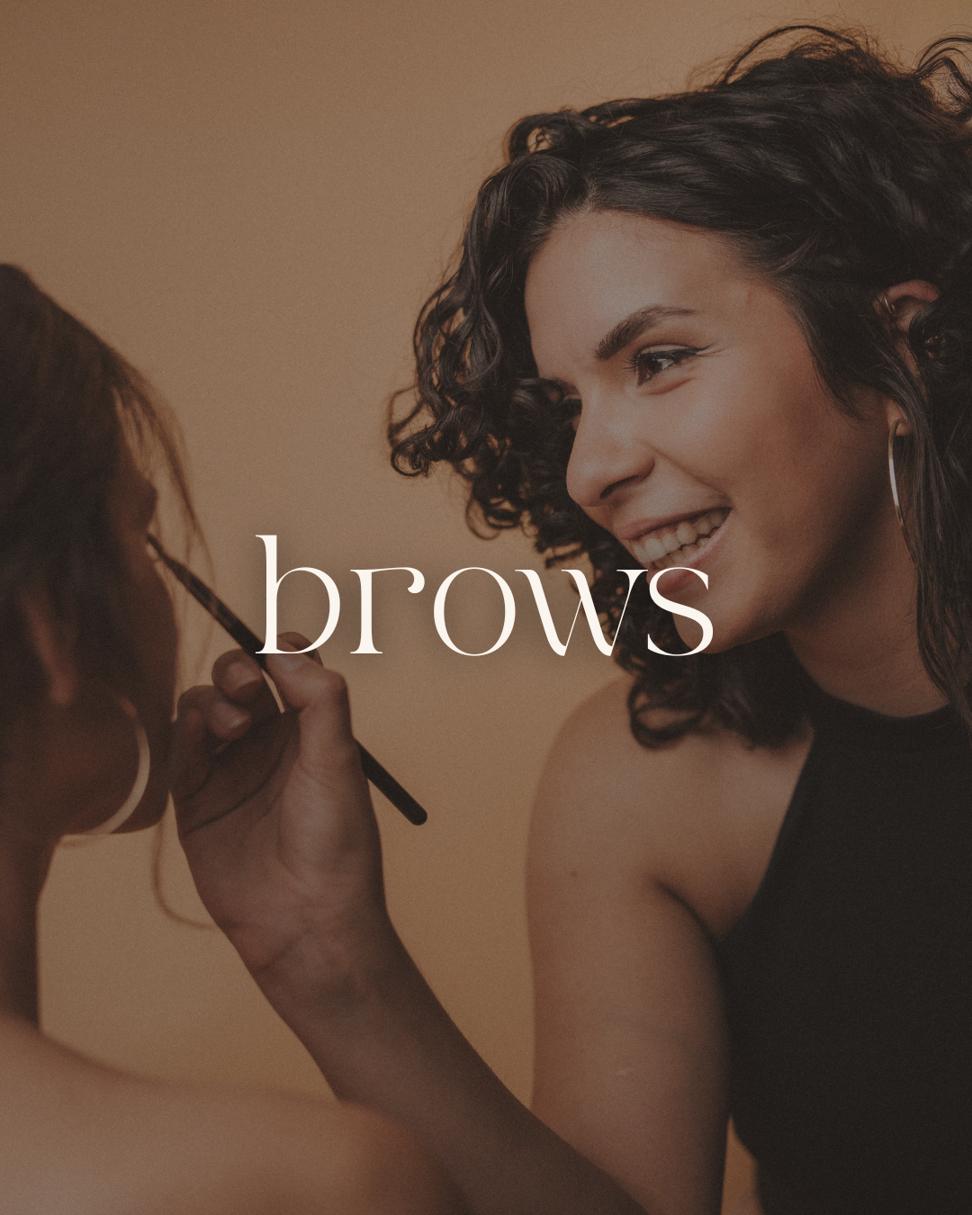 A woman with curly hair smiling as makeup artist applies eyebrow makeup, with the word 'brows' overlayed in the center.