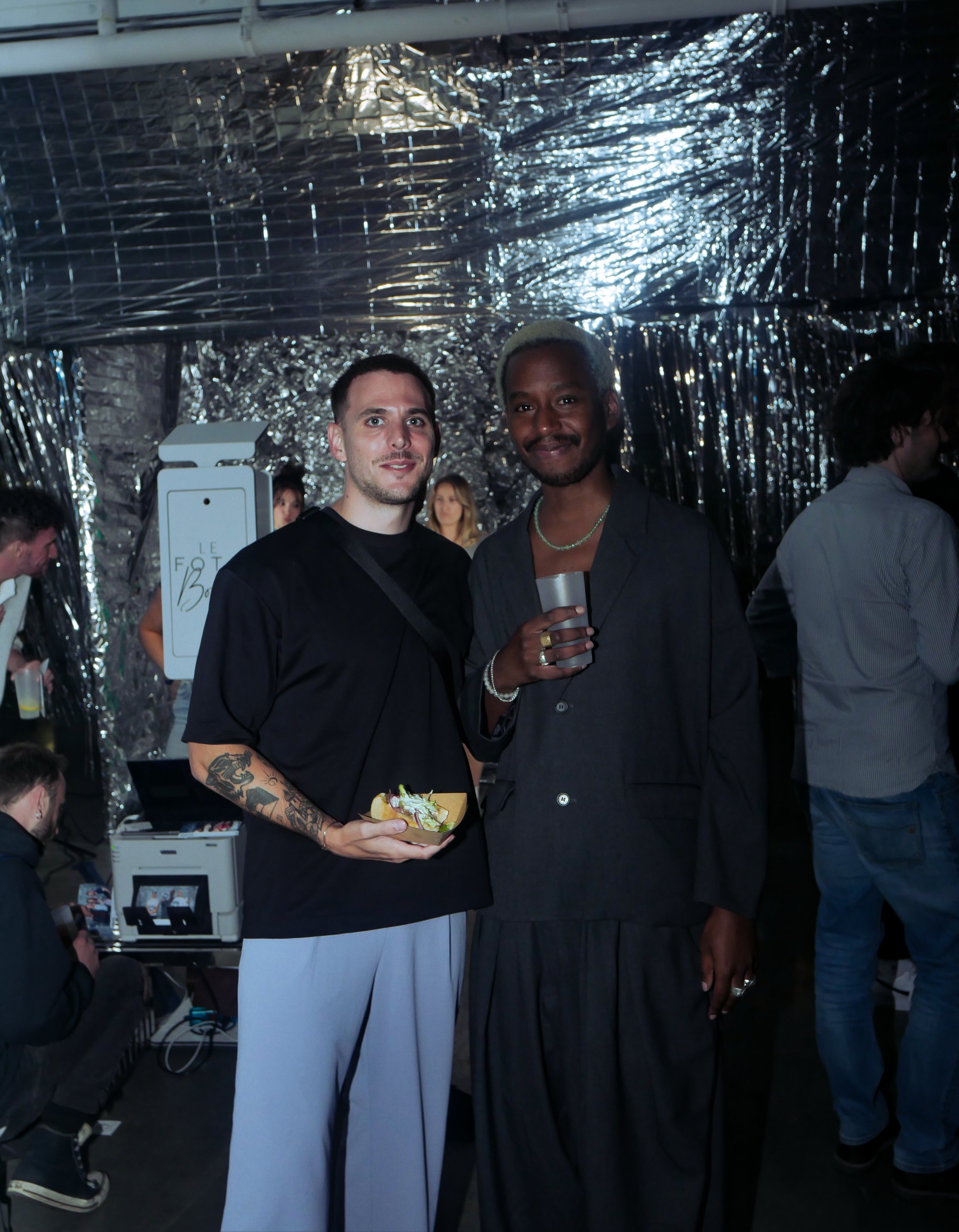 Two men standing together at an event, one holding a plate of food and the other holding a drink, with a shiny silver foil backdrop and other people in the background.