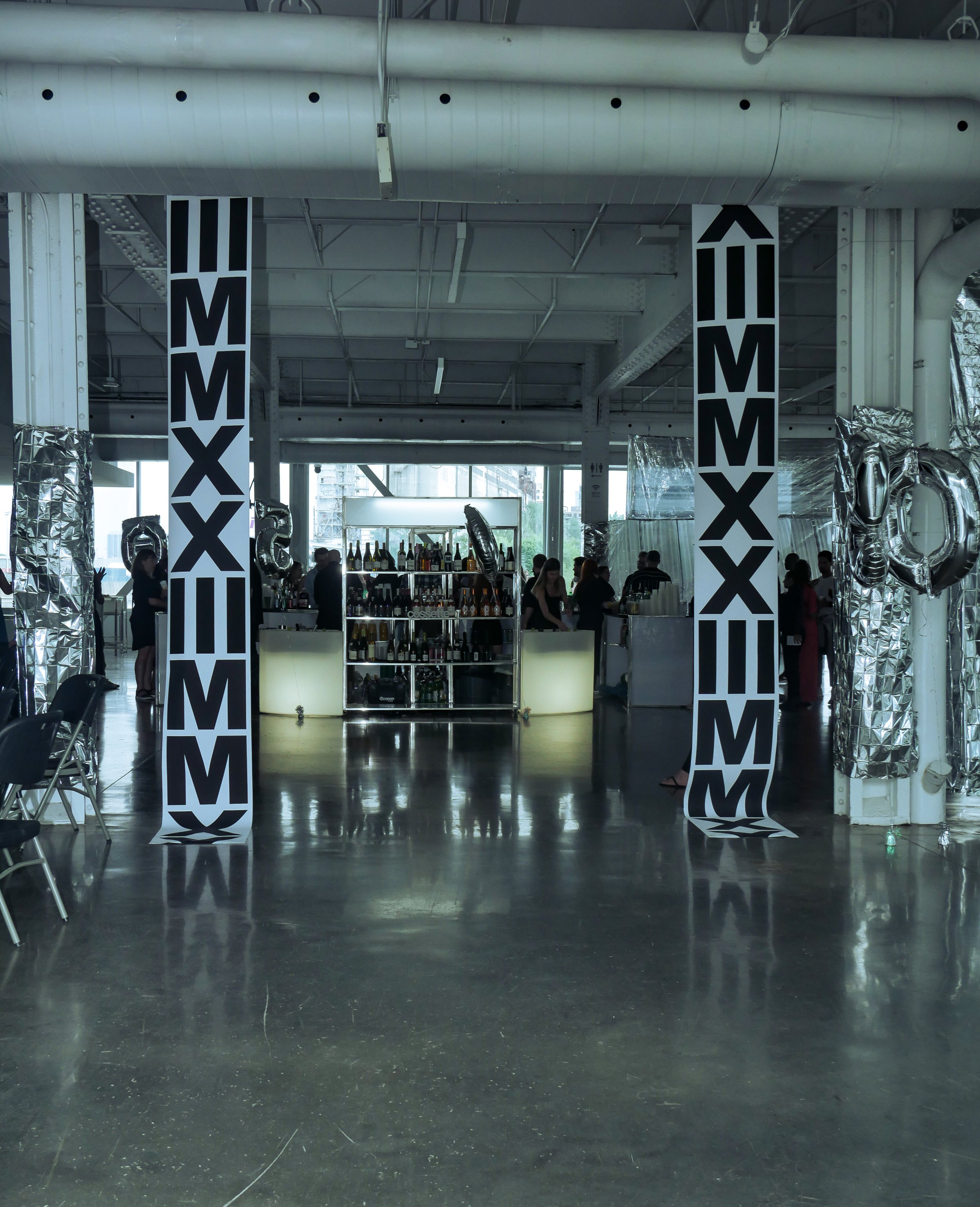 Interior of a modern event space with two large black and white banners that read 'M M X X' flanking a central bar area with bottles and glasses, with some silver balloons on the sides.
