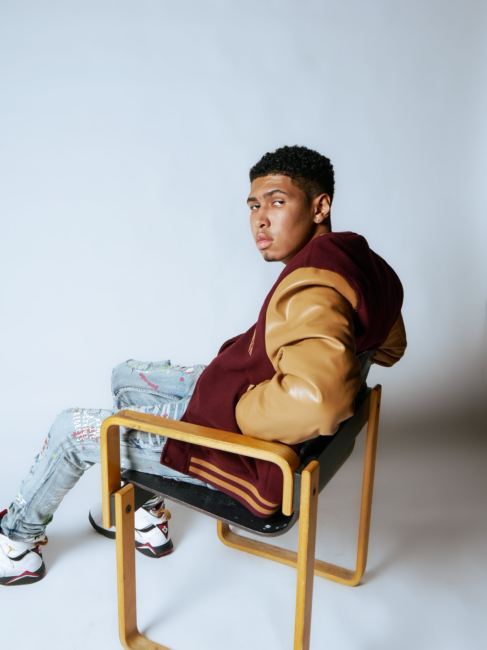 Young man sitting sideways in a black and wooden chair against a plain white background, wearing a maroon and tan hoodie, distressed jeans, and sneakers.