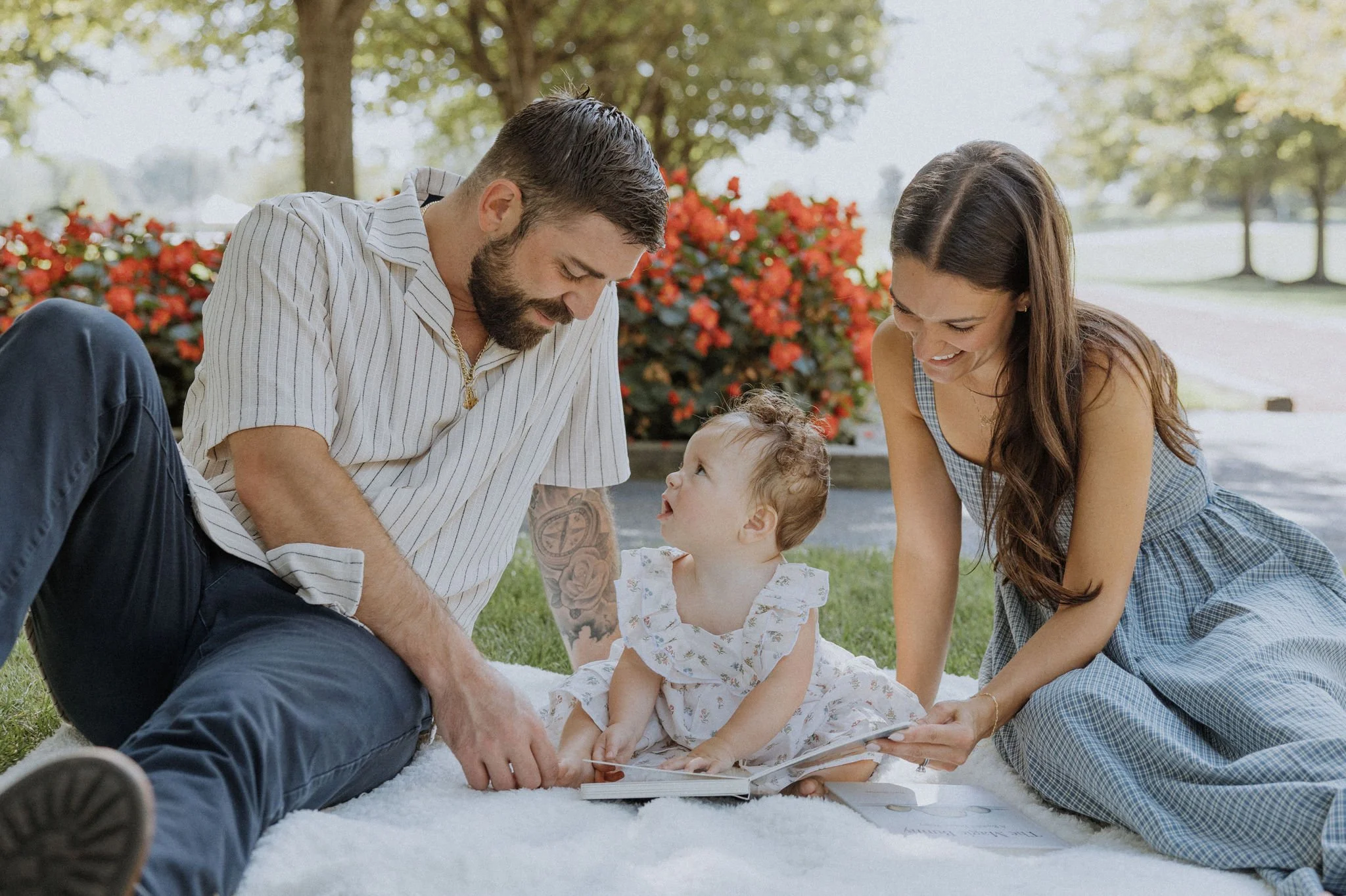 A family of three sitting on a white blanket outdoors, mommy and daddy looking at their young daughter with a book, surrounded by flowers and trees.