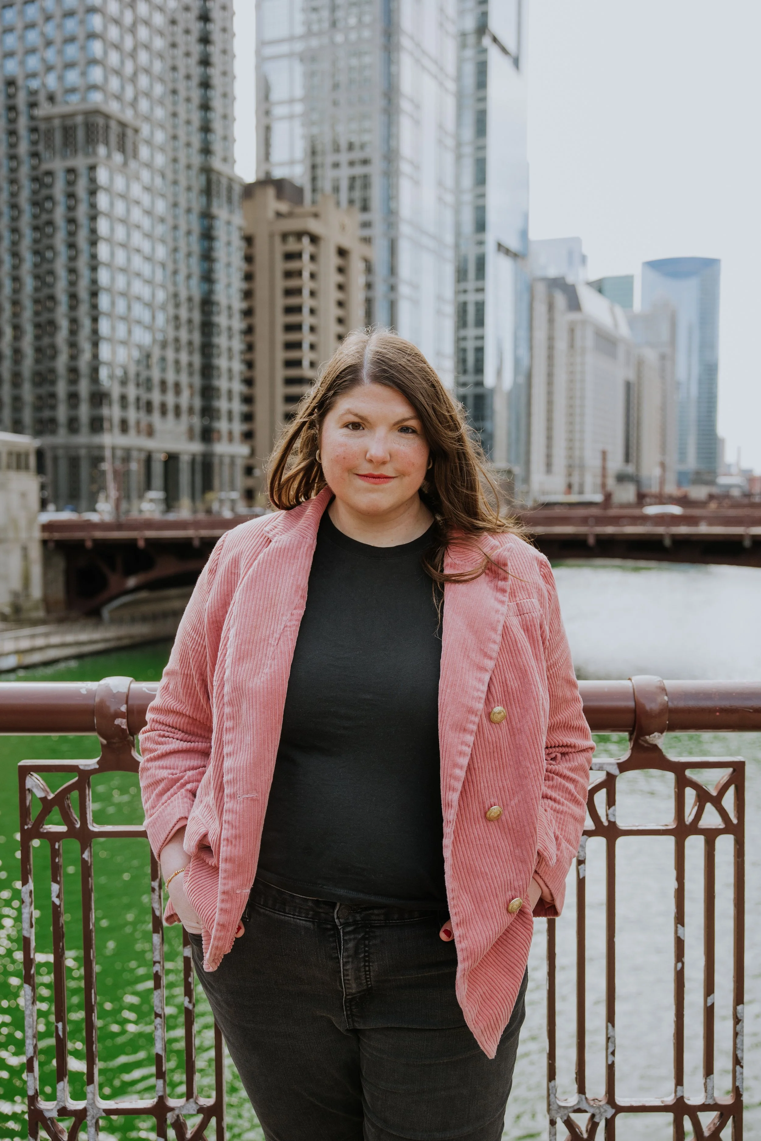 A woman with long brown hair wearing a pink corduroy blazer and black shirt standing on a bridge over a river, with modern skyscrapers in the background.