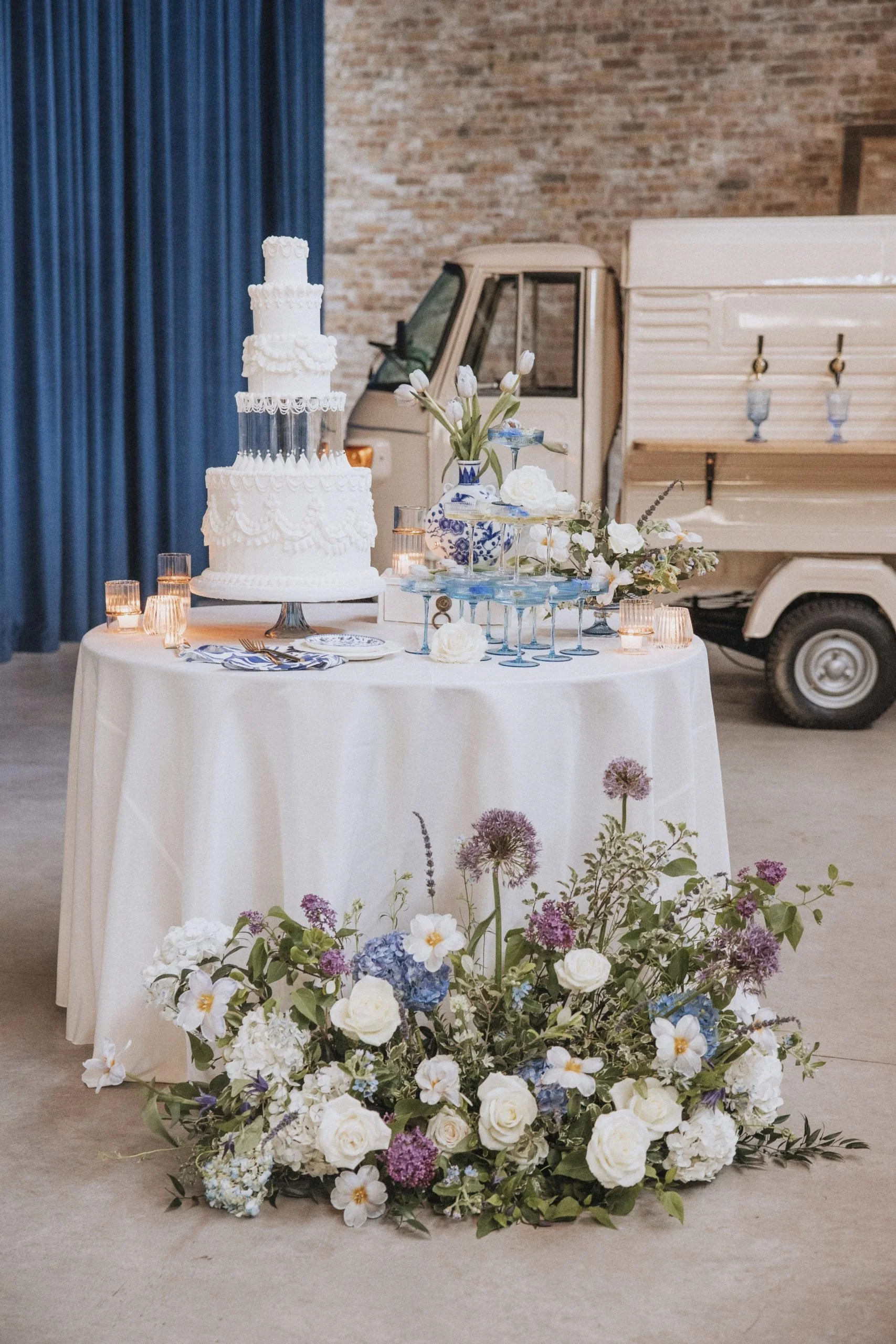 Elegant wedding cake on a round table decorated with white and purple flowers, ivory tablecloth, and small candles, with a vintage vehicle and brick wall in the background.
