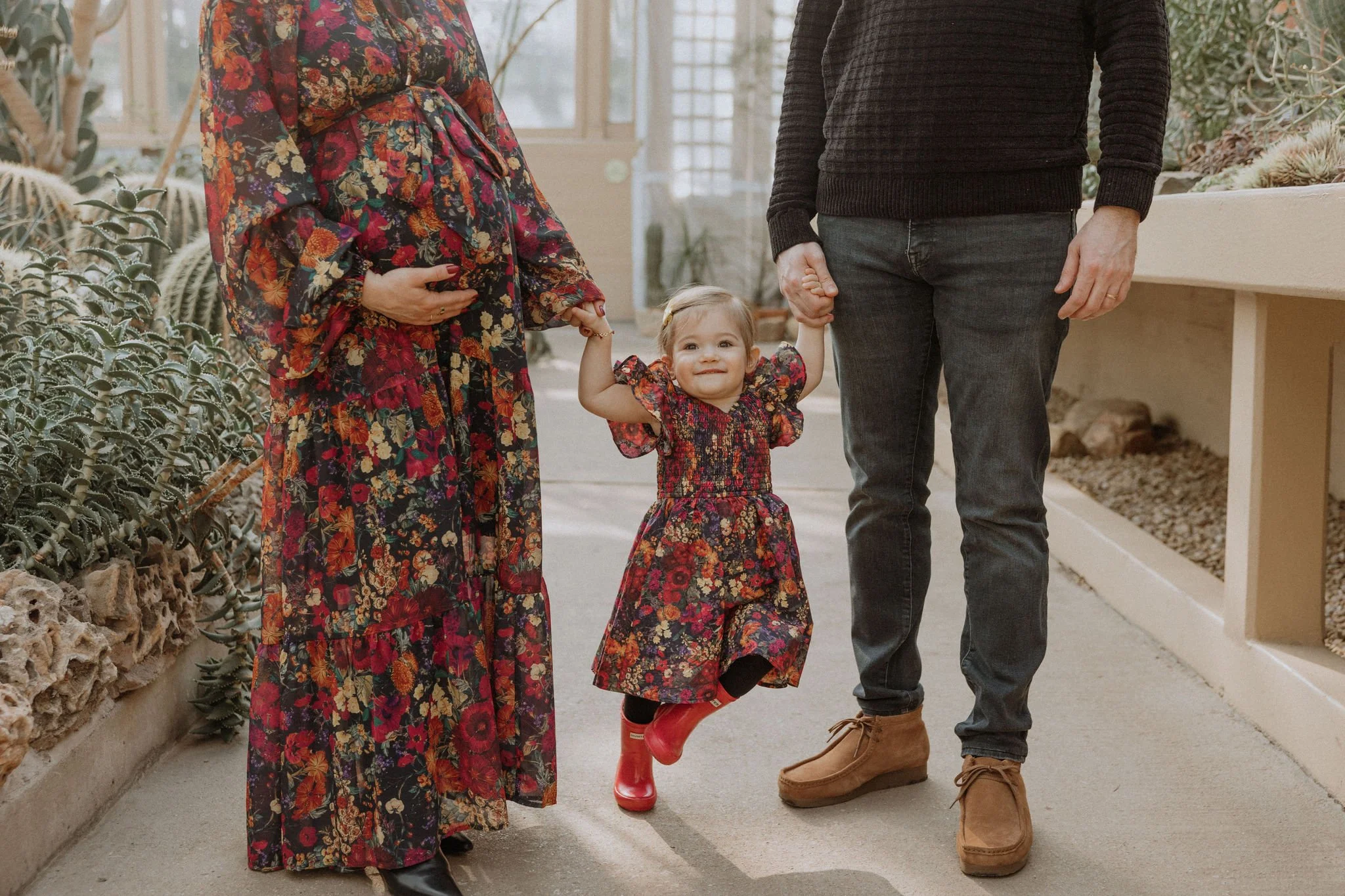 A toddler girl in a floral dress and red rain boots holding hands with two adults, possibly her parents, walking indoors in a greenhouse or conservatory with plants and a tiled pathway.