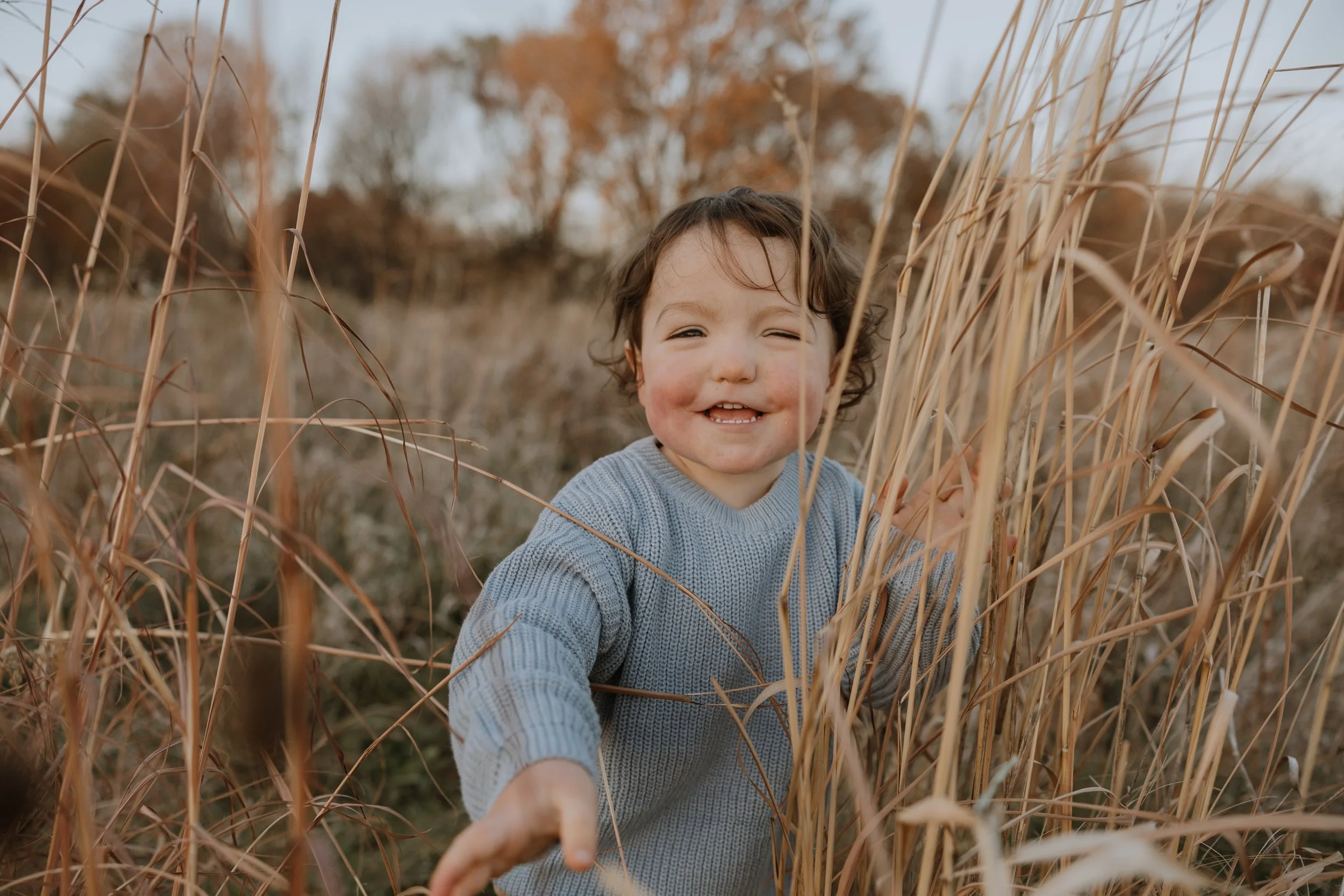 A smiling child with curly hair, wearing a light blue sweater, playing in tall dry grass outdoors during autumn.