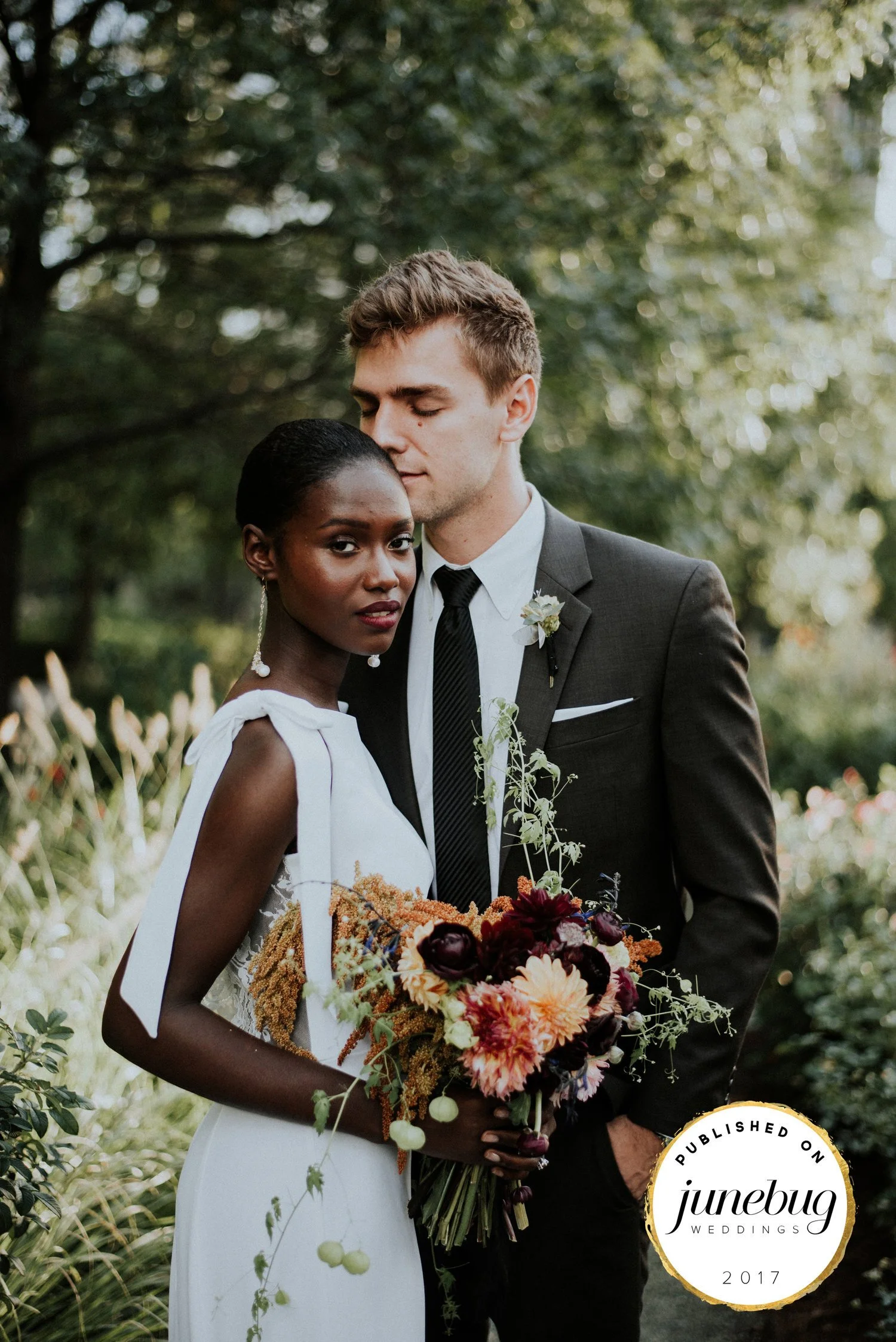 A young couple in wedding attire standing closely outdoors, holding a bouquet of flowers, with trees and greenery in the background.