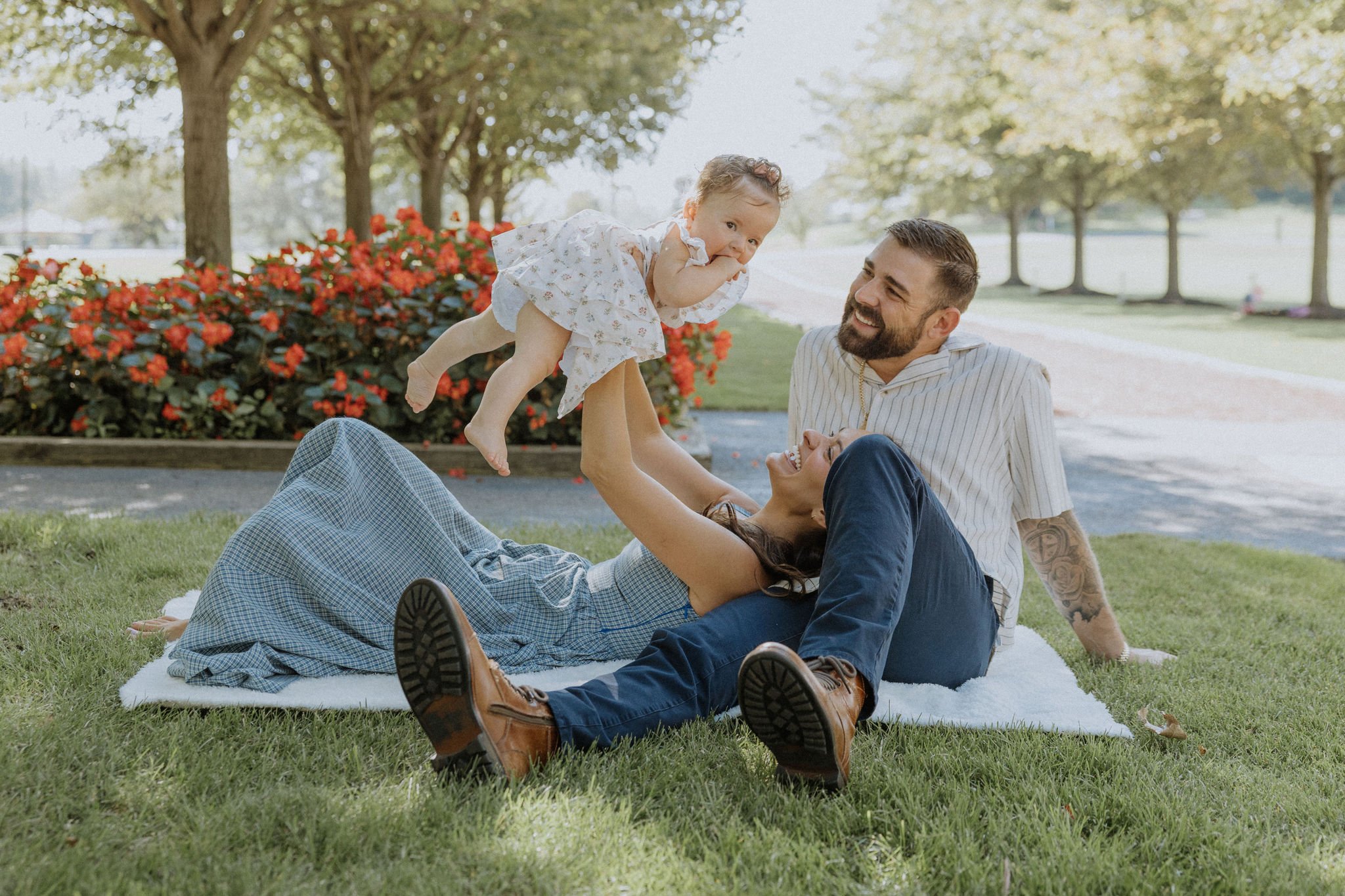 Family of three playing on the grass in a park, with a young child lifted by her mother and the father sitting beside them, all smiling and enjoying a sunny day.