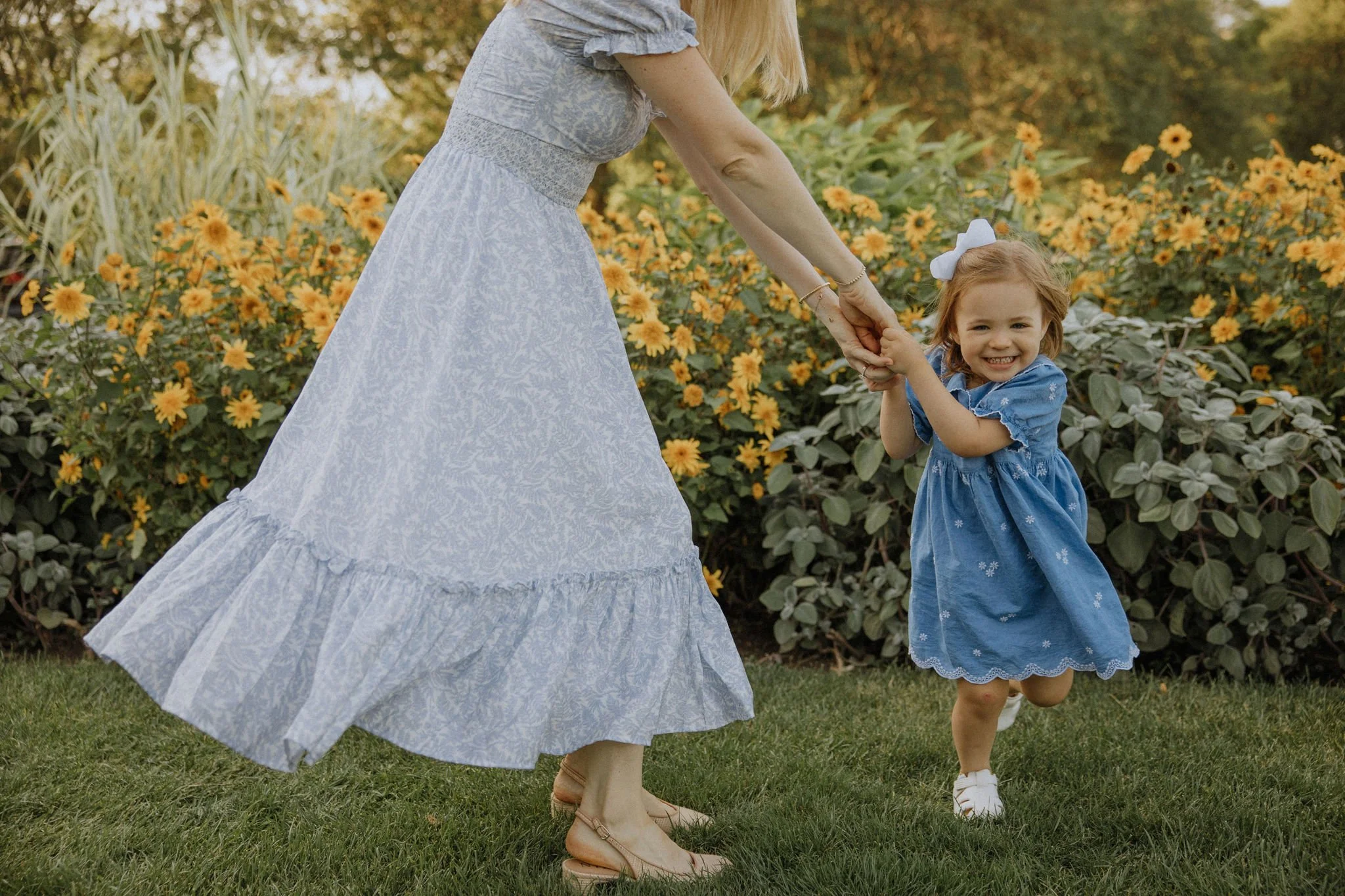 A young girl in a blue dress with a white bow in her hair holding hands and smiling with an adult woman in a long, flowing dress outdoors surrounded by yellow flowers and greenery.