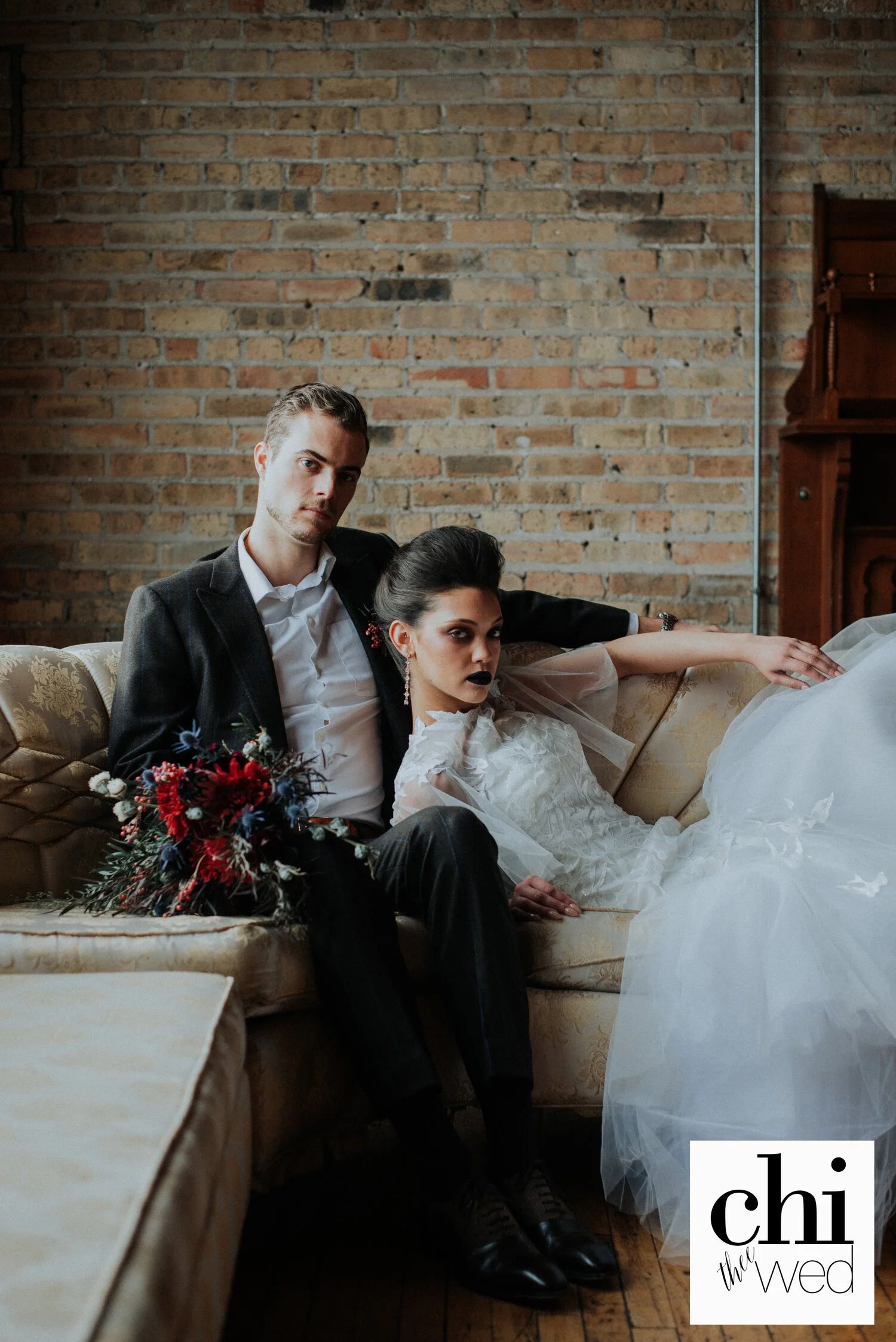 A man and woman, both dressed in wedding attire, sitting on a vintage sofa with a exposed brick wall in the background. The woman is wearing a white wedding dress with dark lipstick, and the man is in a dark suit holding a bouquet of red and blue flowers.