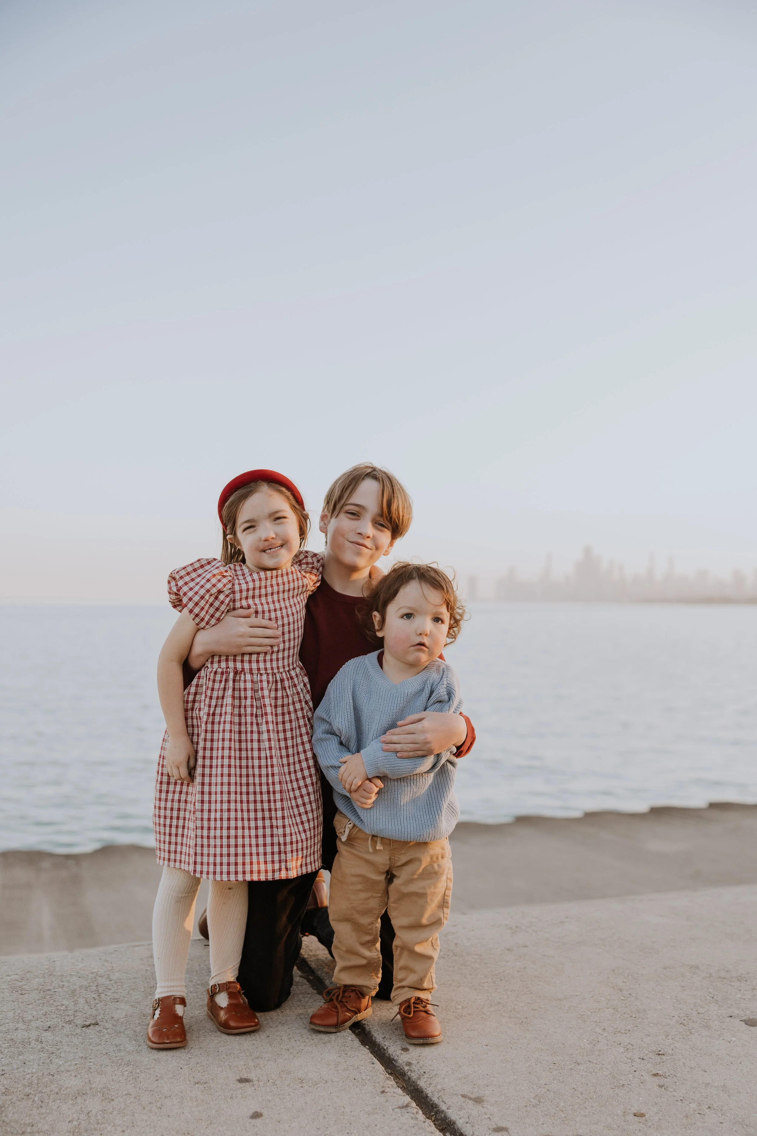 Three children, a girl and two boys, standing together near the water's edge with a city skyline in the background, during sunset.