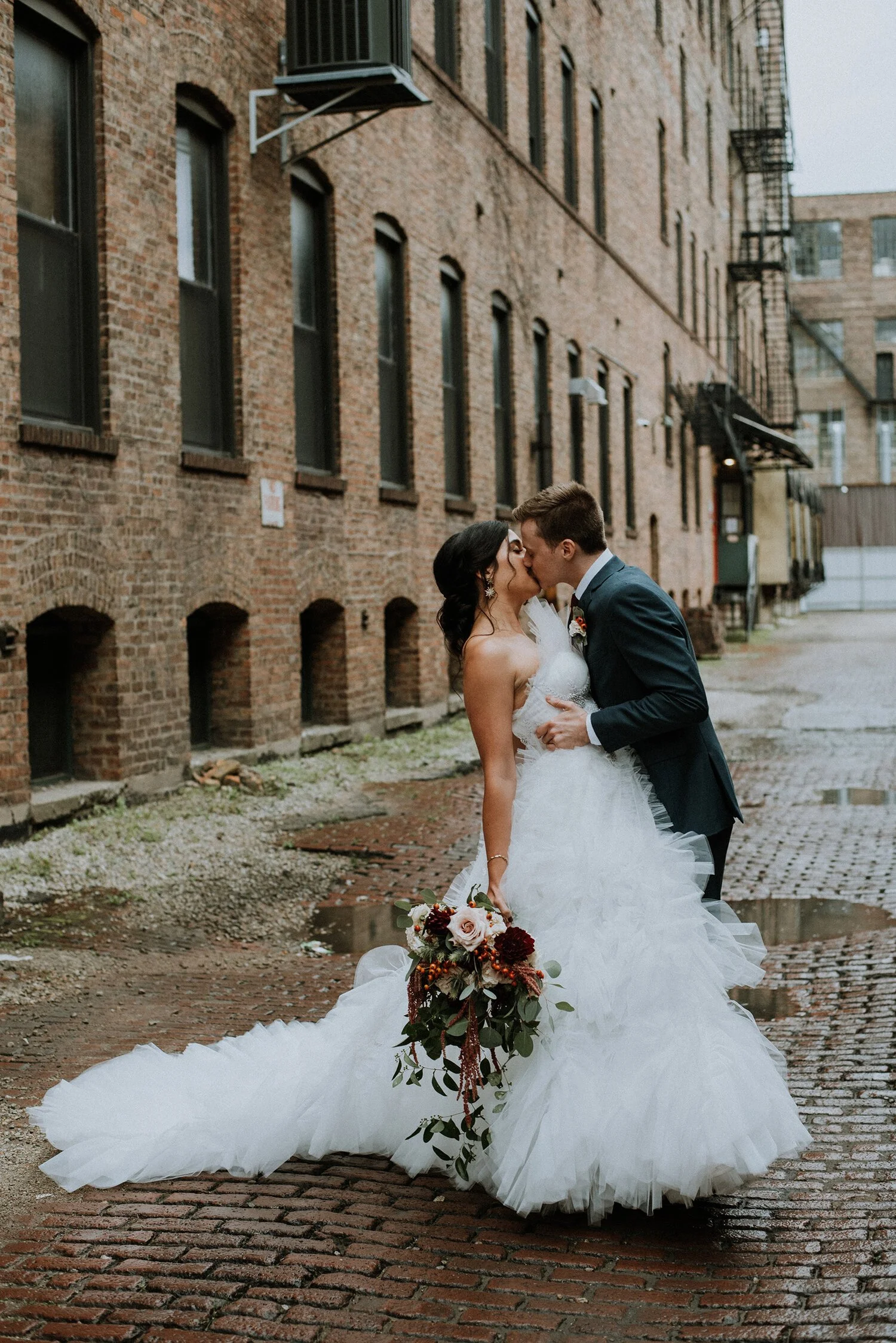 Bride and groom kiss on a brick alley, with the bride holding a bouquet of flowers.