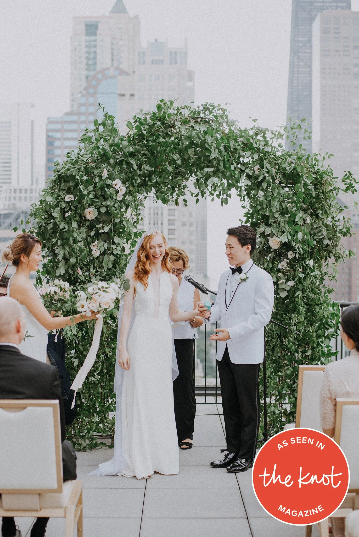 A wedding ceremony on a rooftop with a green floral arch, a bride in a white gown, a groom in a white tuxedo jacket, and a crowd of guests in an urban setting.