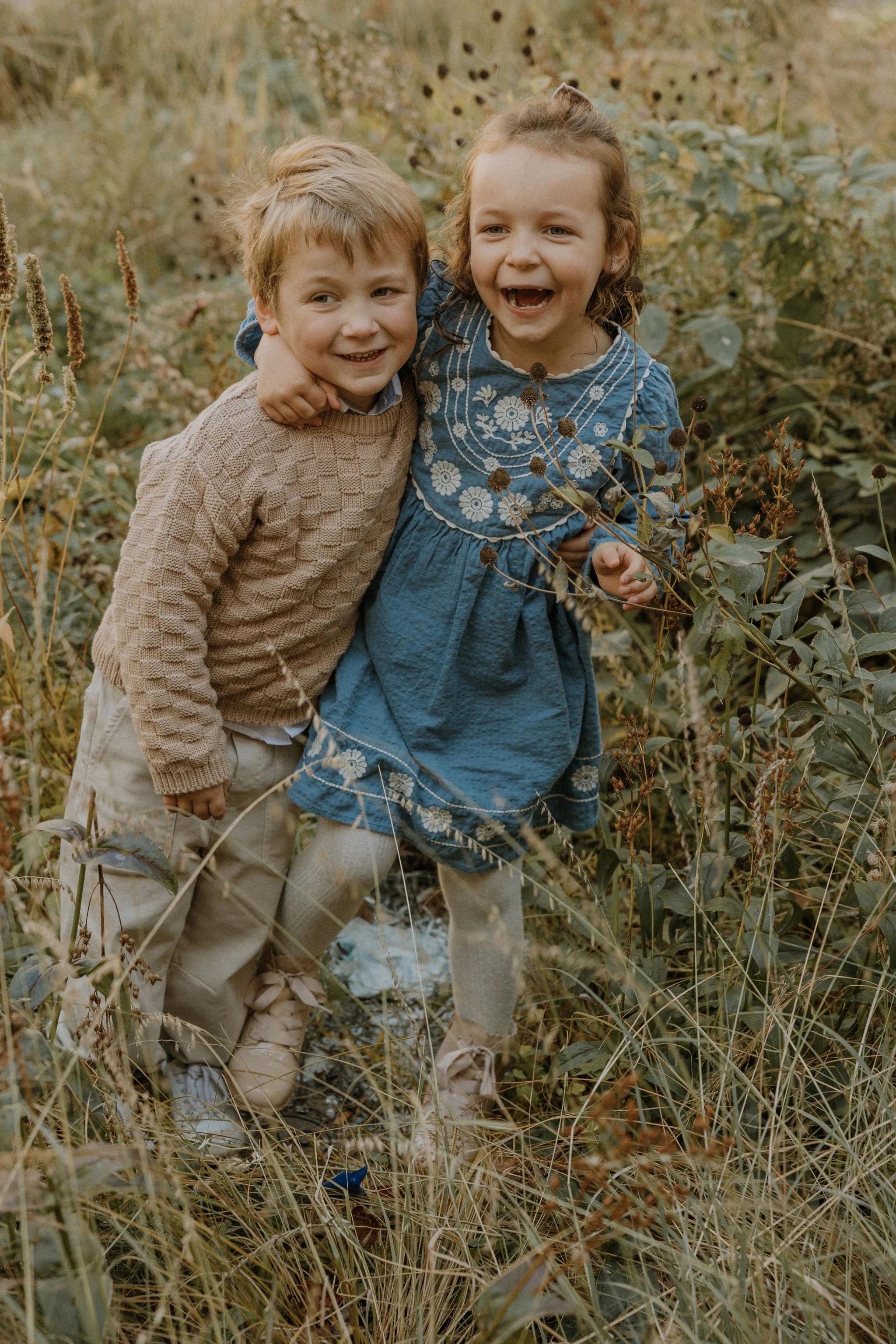 Two children, a boy and a girl, happily hugging outdoors in a natural, grassy setting with surrounded foliage.