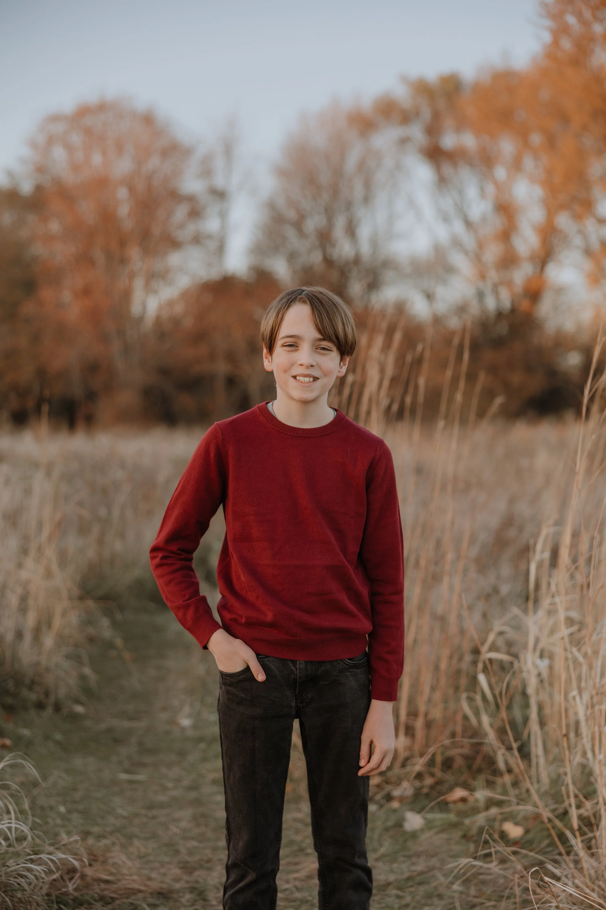 A young boy with brown hair and a red sweater standing outdoors on a path surrounded by tall dry grasses and autumn trees in the background.