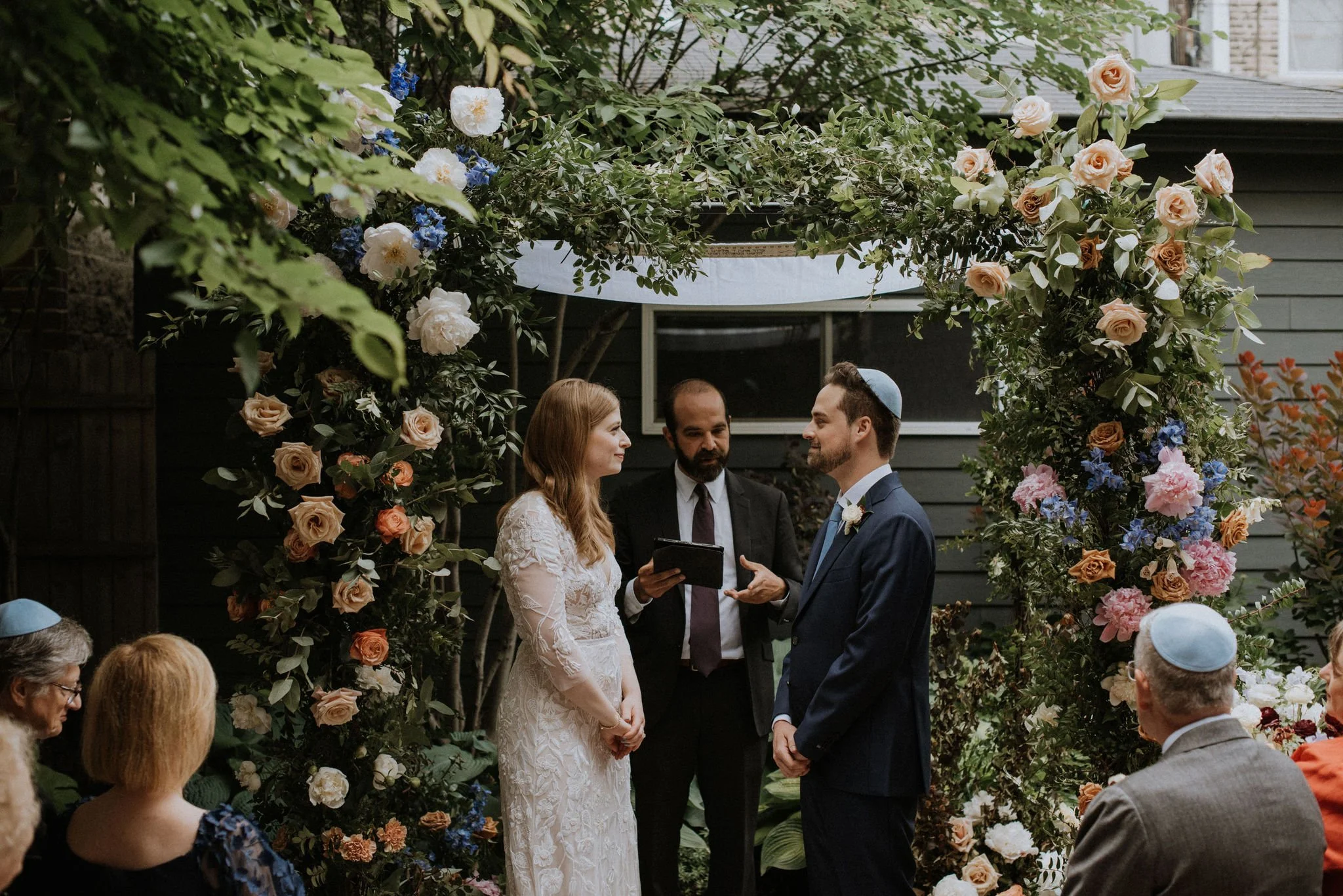 A couple getting married in an outdoor ceremony under a floral arch, with guests sitting nearby and officiant reading vows.