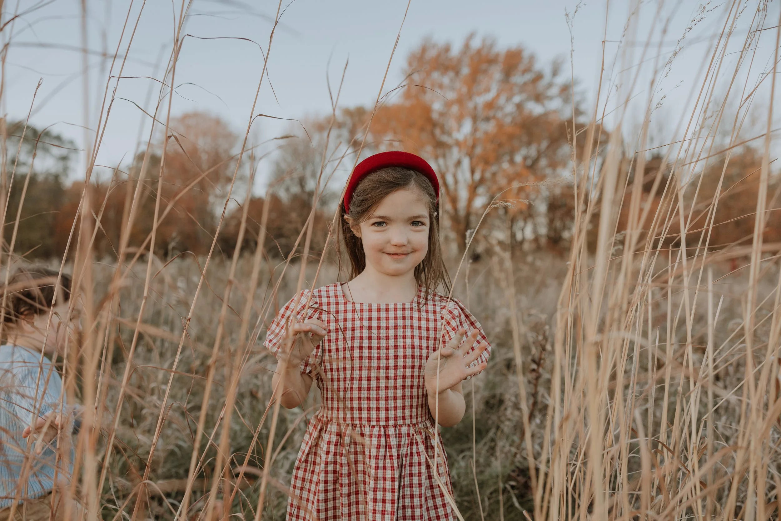 A young girl with light skin, brown hair, wearing a red and white checkered dress and a red headband, standing in a field of tall dry grass with trees in the background during autumn.