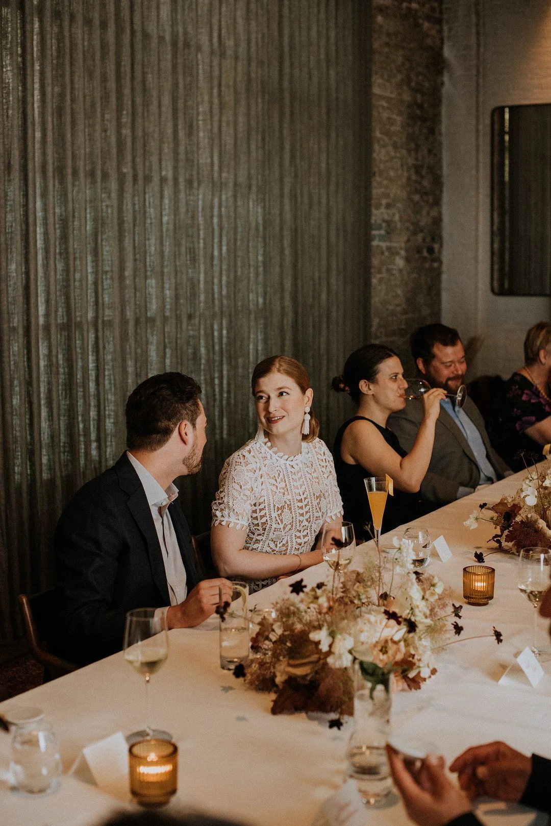 People sitting at a long dining table during a social event, decorated with floral arrangements and candles, with a textured background wall.