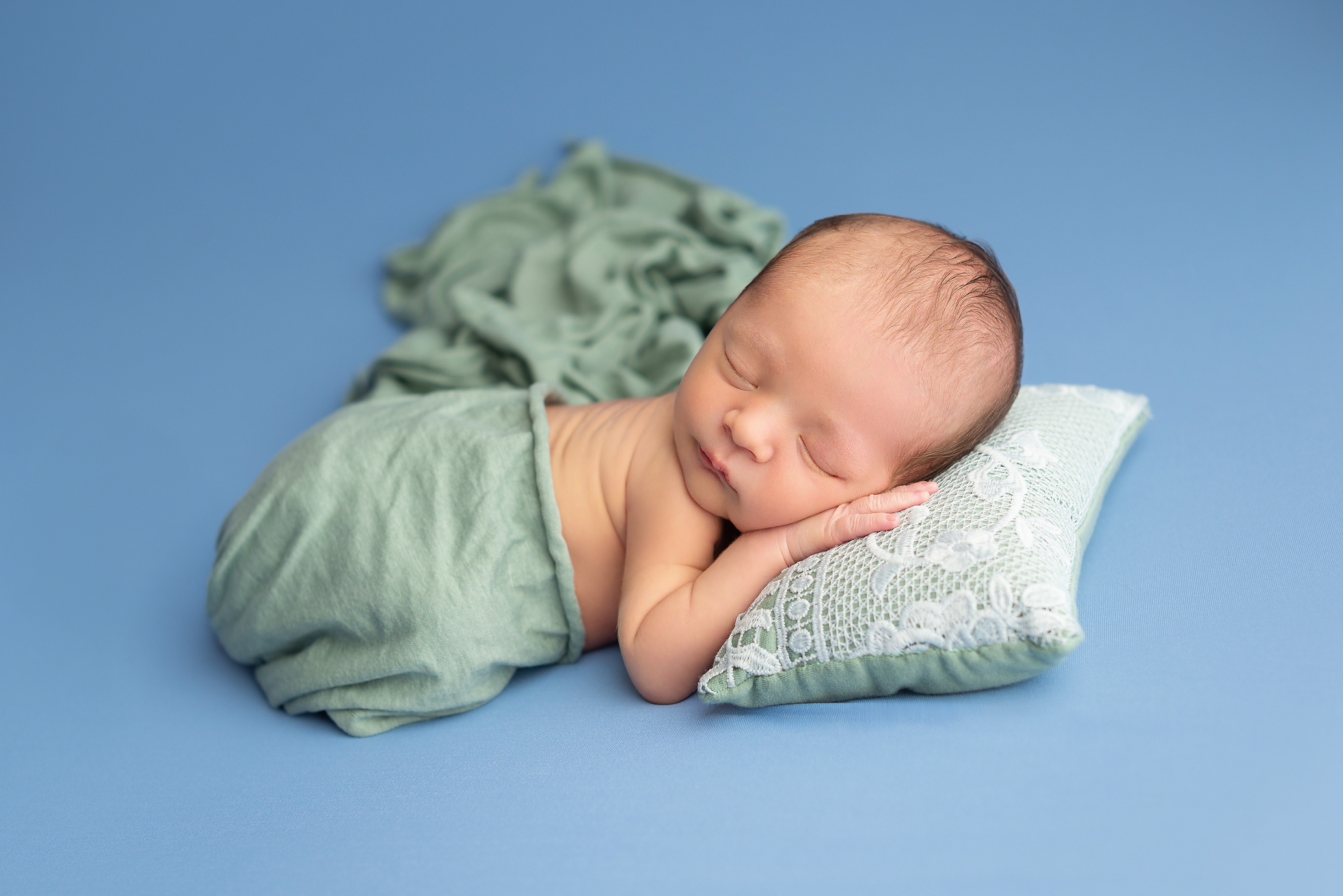 newborn boy laying on blue backdrop with green wrap and pillow