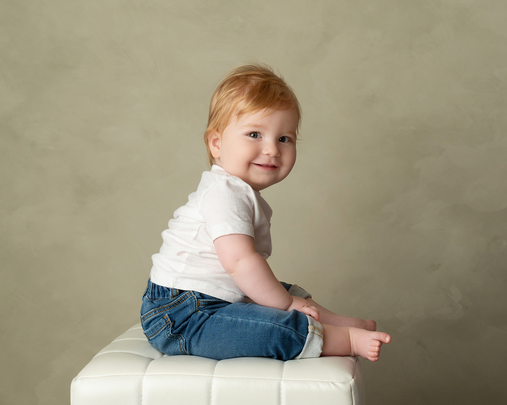 baby boy with red hair sitting on white stool in jeans and white t-shirt