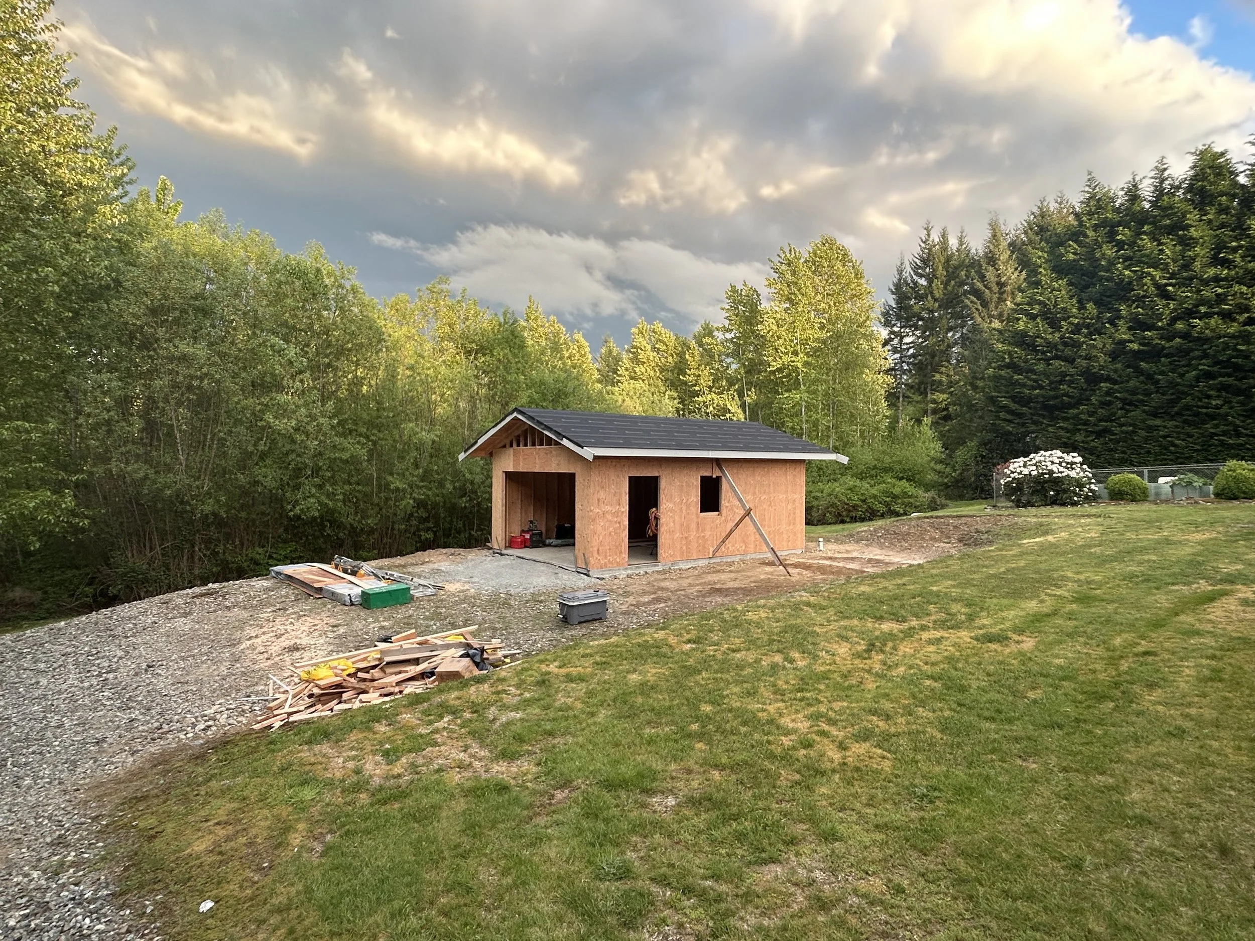 Small wooden shed under construction on a grassy area, with construction materials and tools nearby, surrounded by trees and a partly cloudy sky.