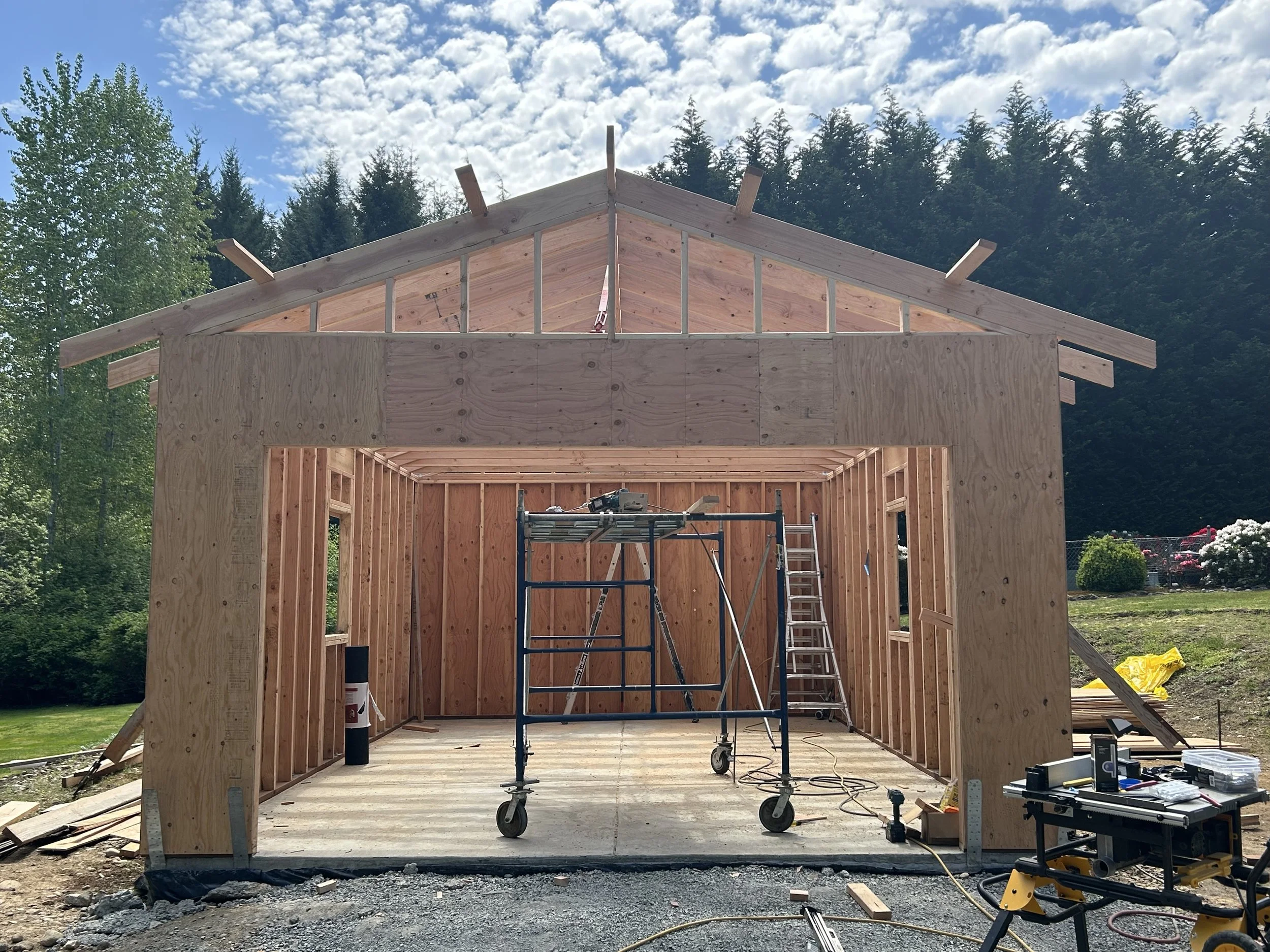 Wooden frame of a house under construction, with scaffolding inside, surrounded by construction tools and materials, under a partly cloudy sky.