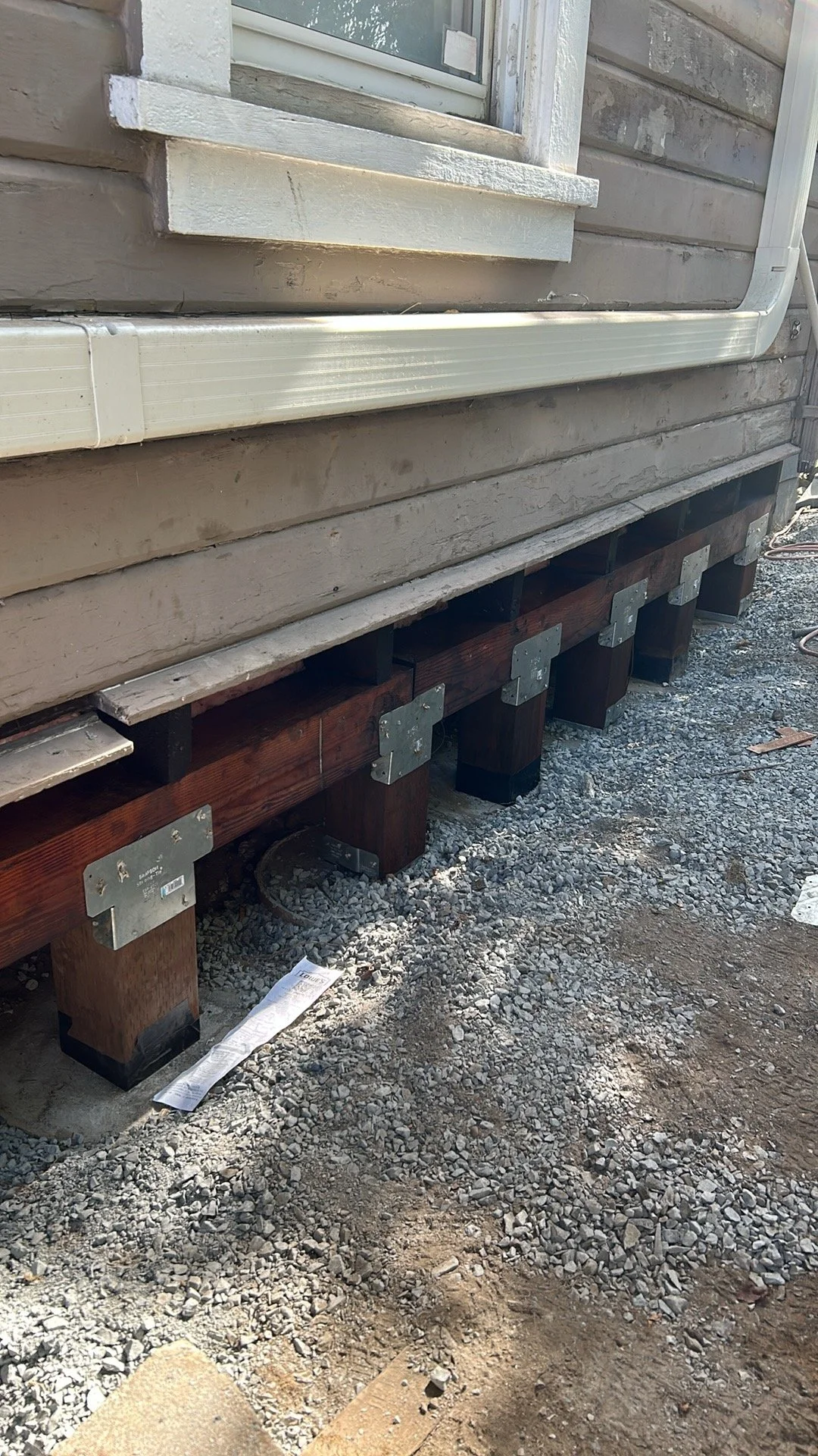 Close-up of a house foundation with wooden support beams and metal brackets, with gravel and dirt in the foreground.