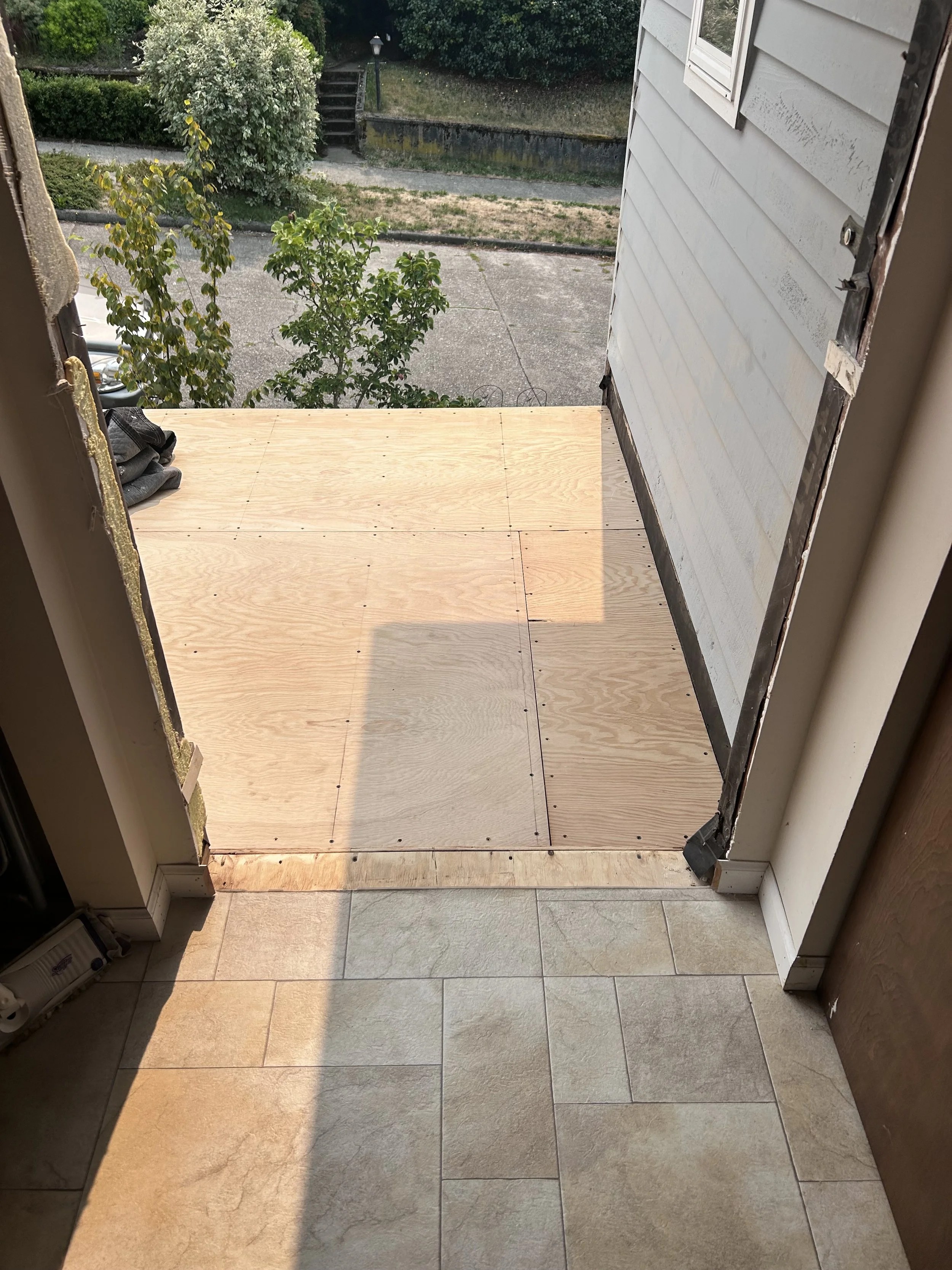 View from inside a house looking out to a small, partially constructed wooden porch with plywood flooring. Outside, there is a concrete sidewalk, some greenery and bushes, a small tree, and a set of stairs leading up to another area. The house's exte