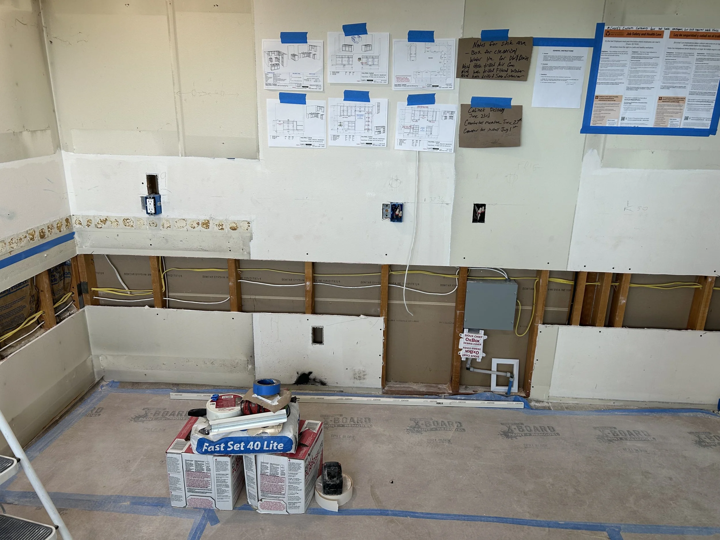 Under-construction room with walls partially finished, electrical wiring exposed, and construction materials on the floor. Blue painter's tape, diagrams on the wall, and a box of fast setting drywall compound present.