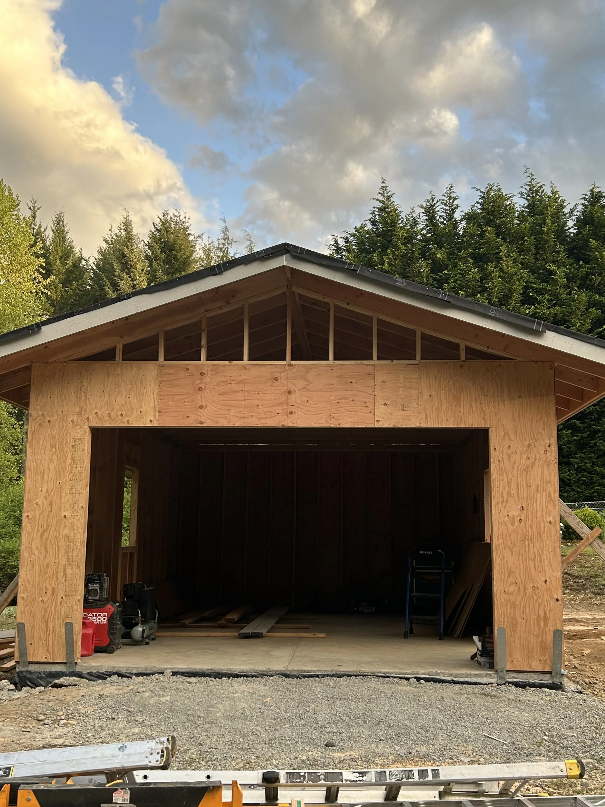 A new wooden garage under construction with a pitched roof, surrounded by trees and a partly cloudy sky.