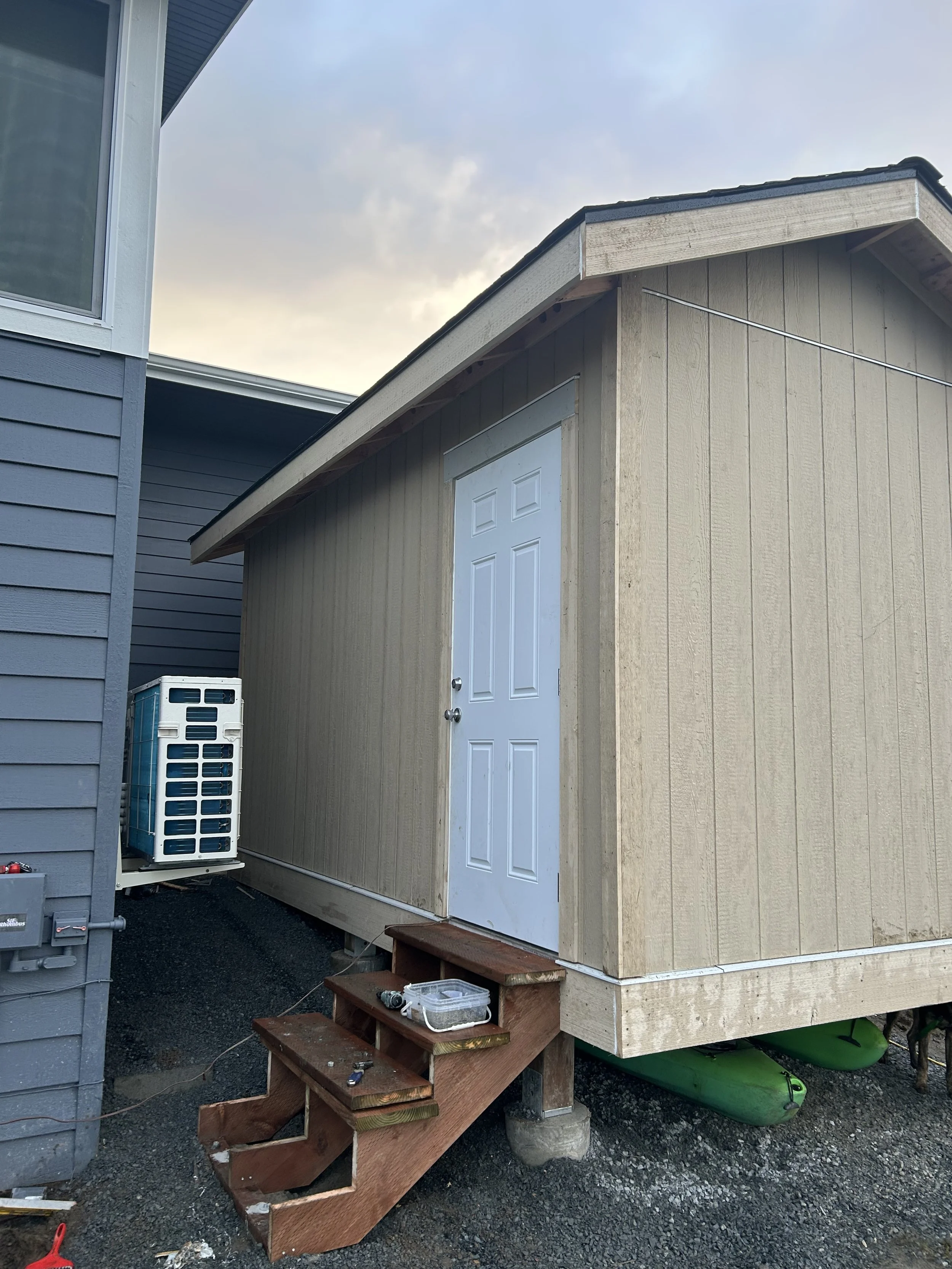 Small beige shed with a white door and wooden steps leading up to it, located between a blue house and an air conditioning unit, with a cloudy sky in the background.