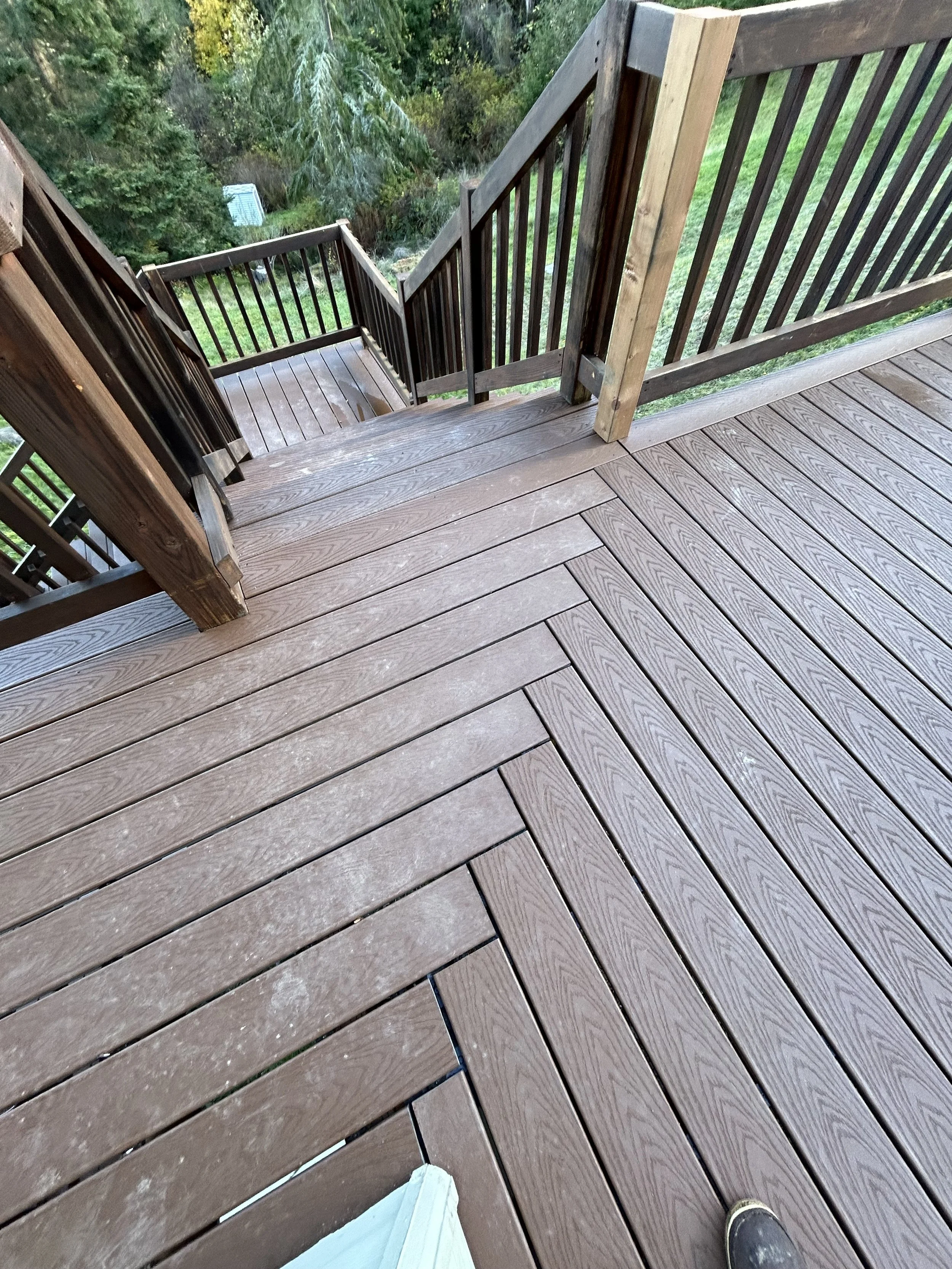 View of a wooden outdoor deck with stairs leading down to a grassy backyard surrounded by trees.