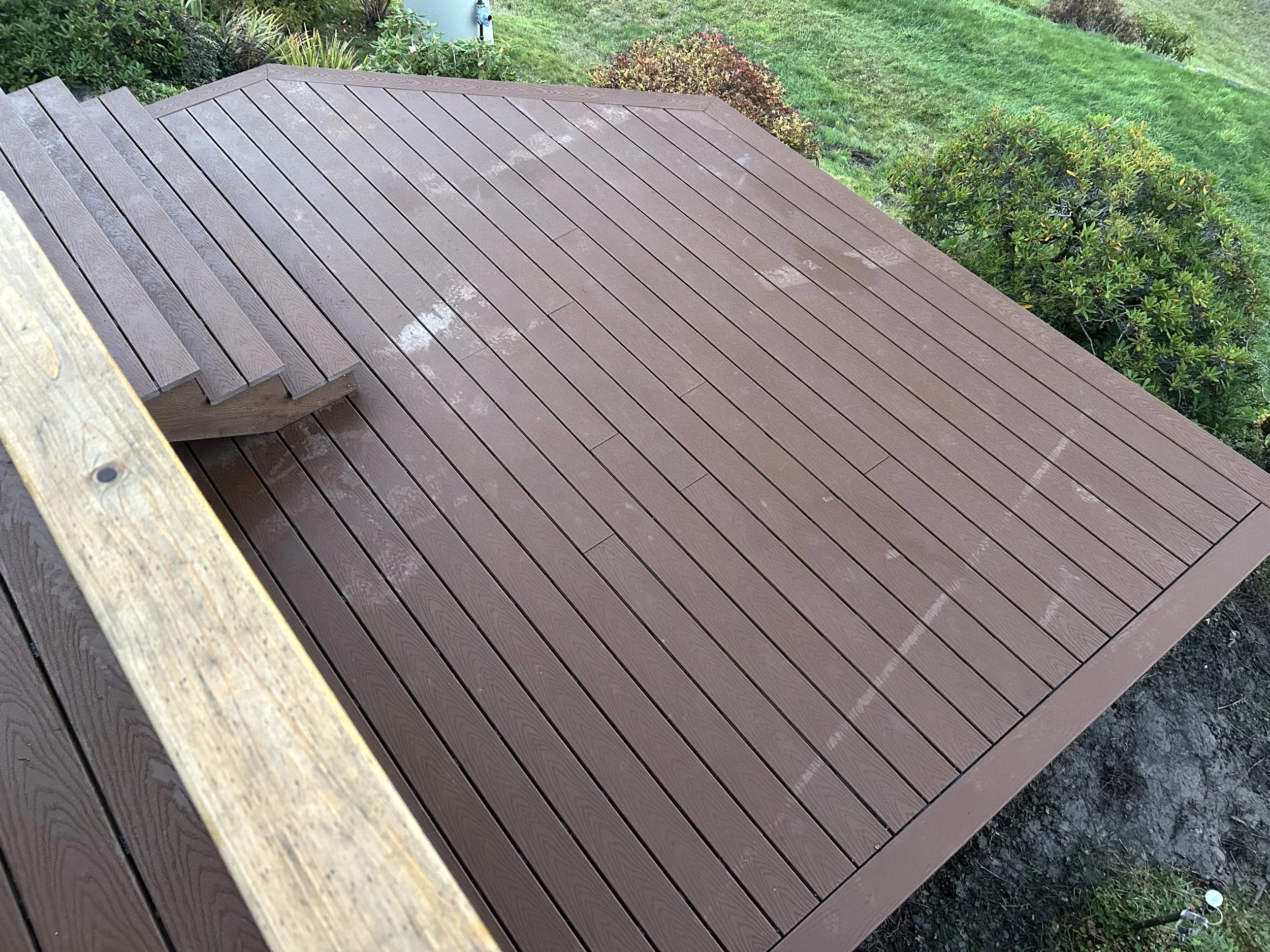 Freshly installed wooden deck with a stair at the corner, surrounded by green grass, bushes, and small plants.