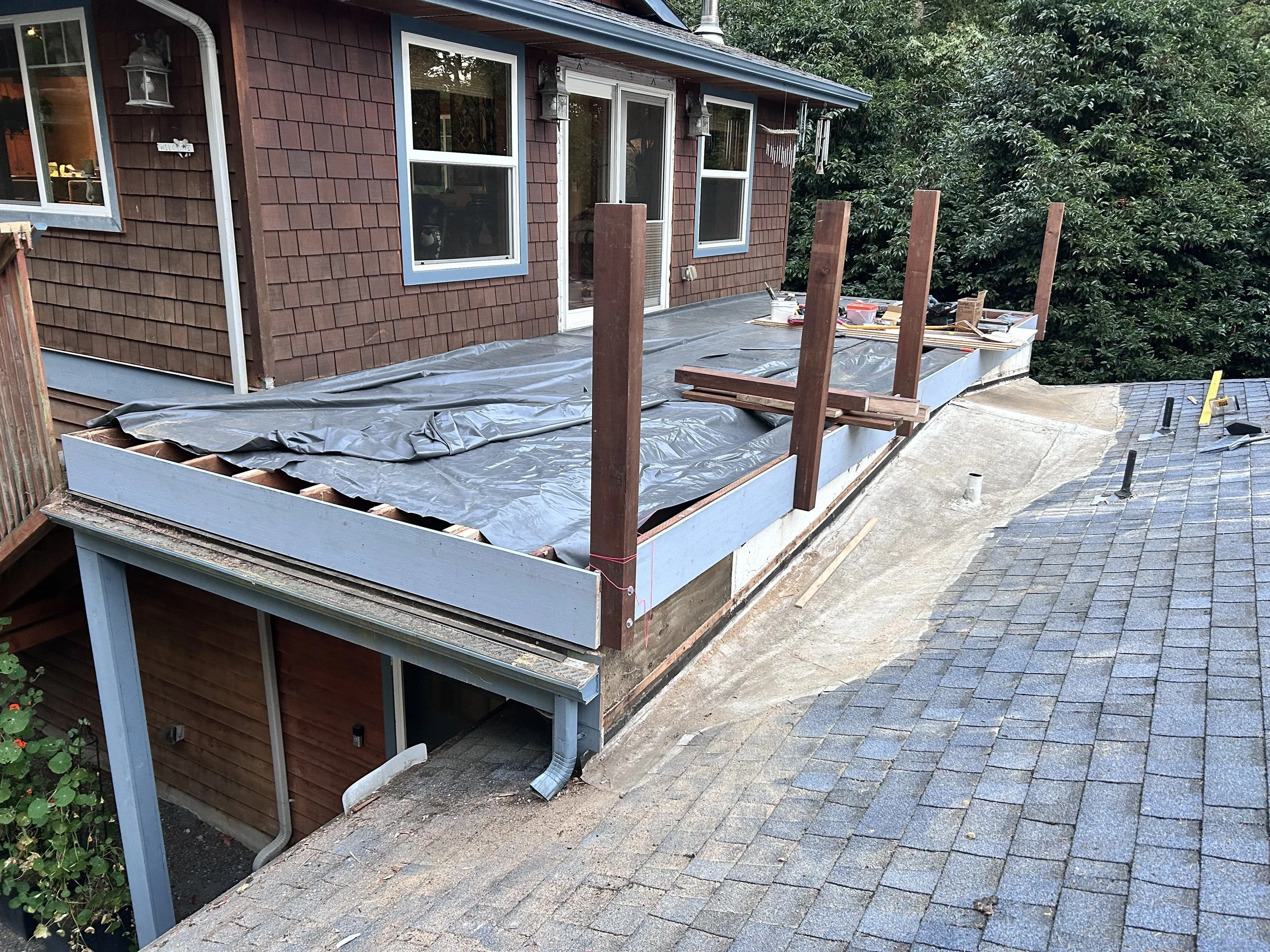 House exterior under construction with a new deck being built, covered with a black tarp, and supported by vertical wooden posts; surrounding the house are trees and a sloped asphalt shingle roof with vents.
