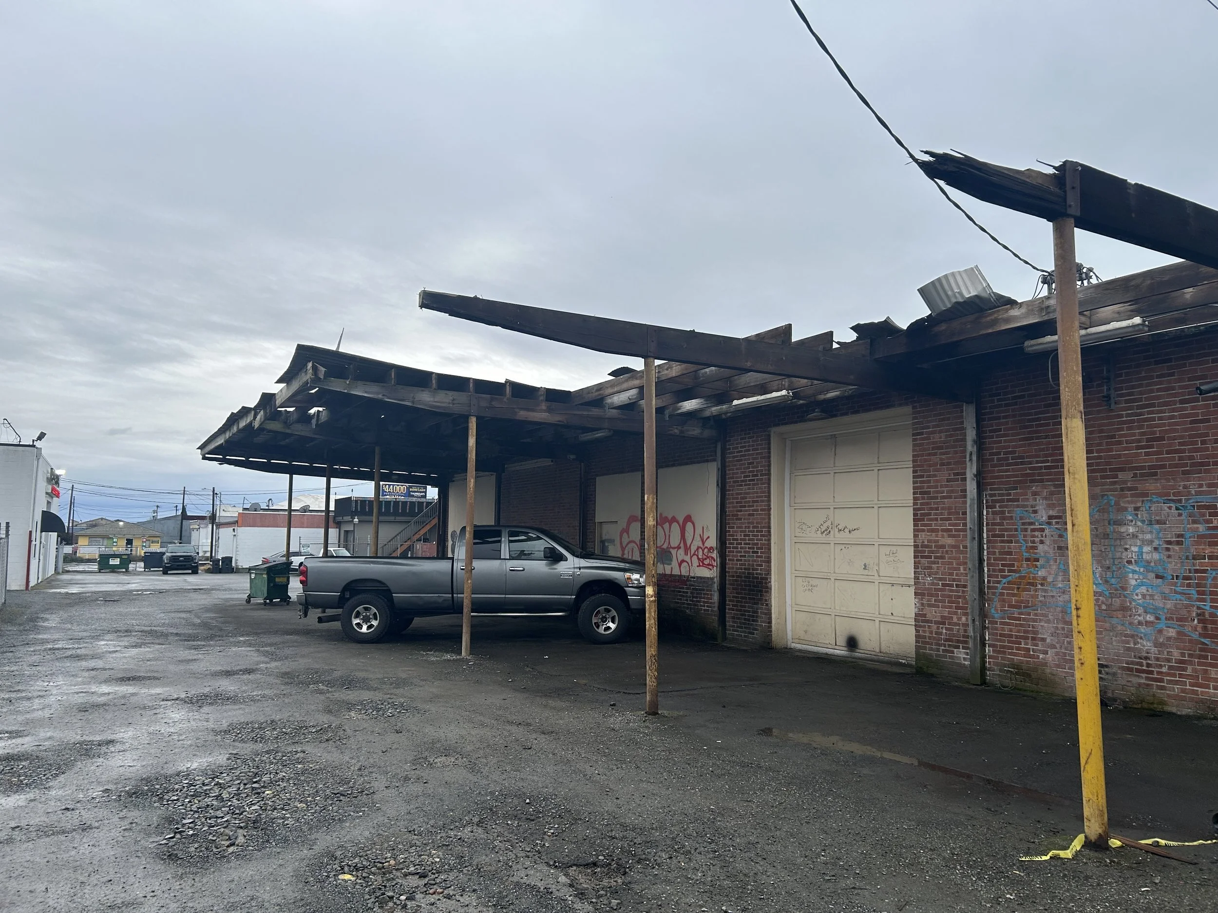 An abandoned building with a damaged roof, graffiti on the brick wall, a silver pickup truck parked underneath, and an unpaved, gravel lot with puddles and scattered debris.