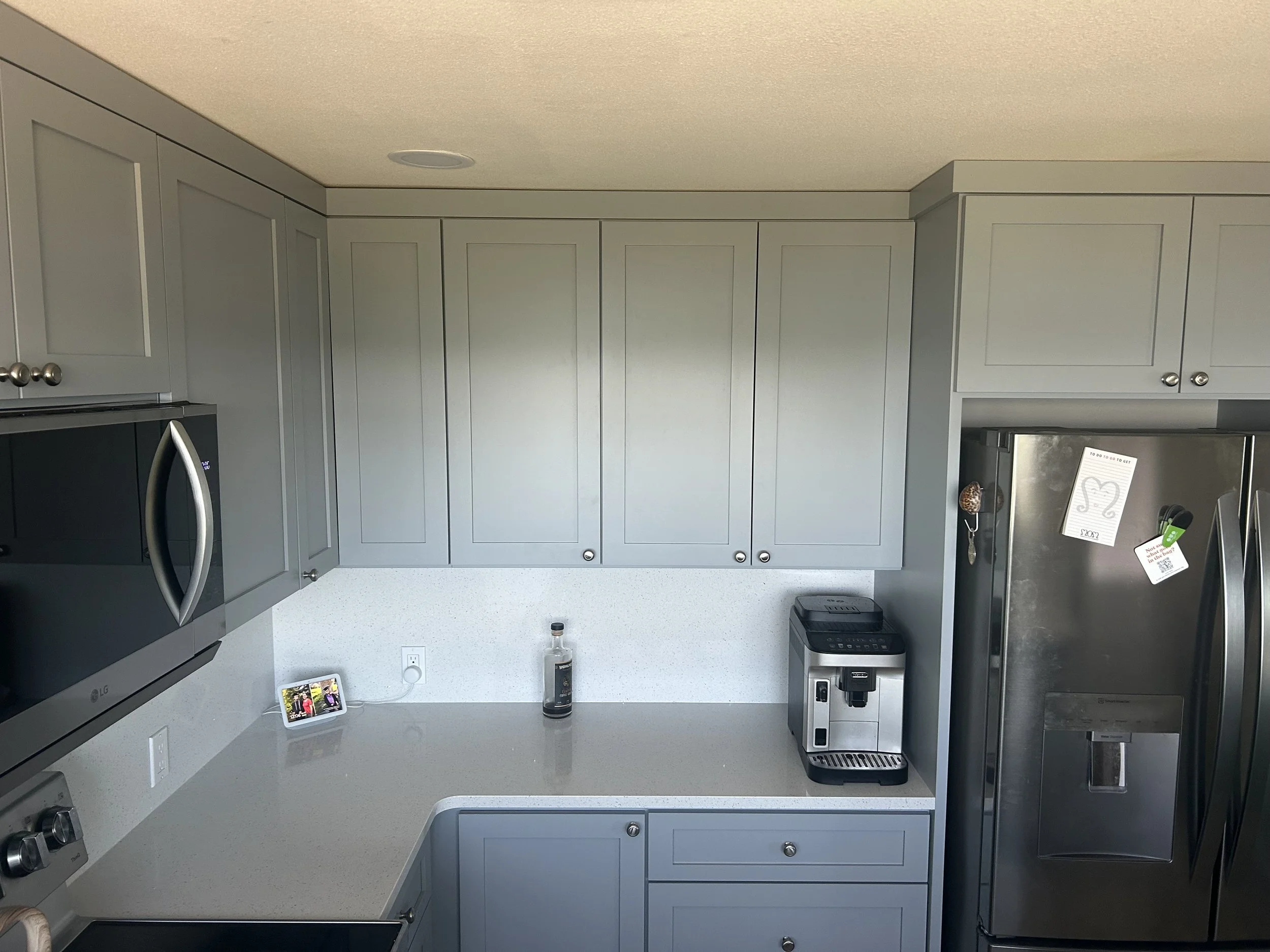 Modern kitchen with gray cabinets, stainless steel refrigerator, microwave oven, coffee machine, and a small smart display device on the countertop, with a bottle and some keys on the counter.