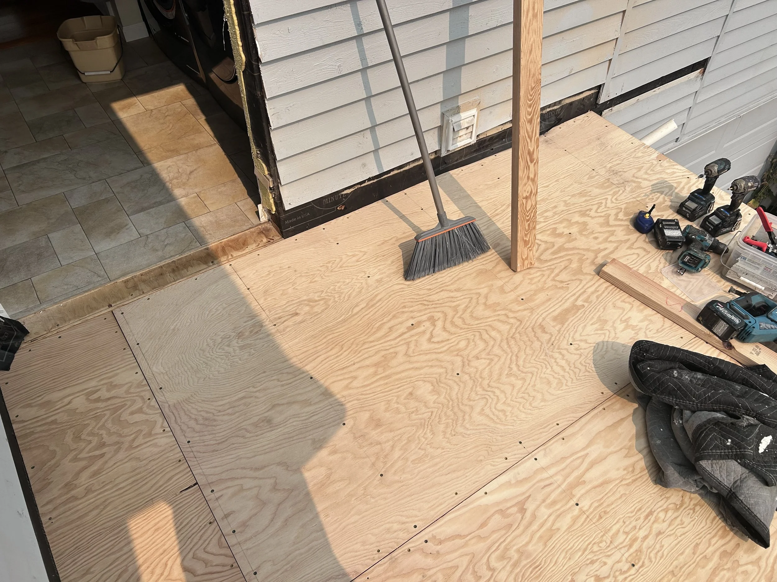 Plywood flooring being installed outside a house, with construction tools, cordless drills, a toolbox, a broom, and a work jacket visible.