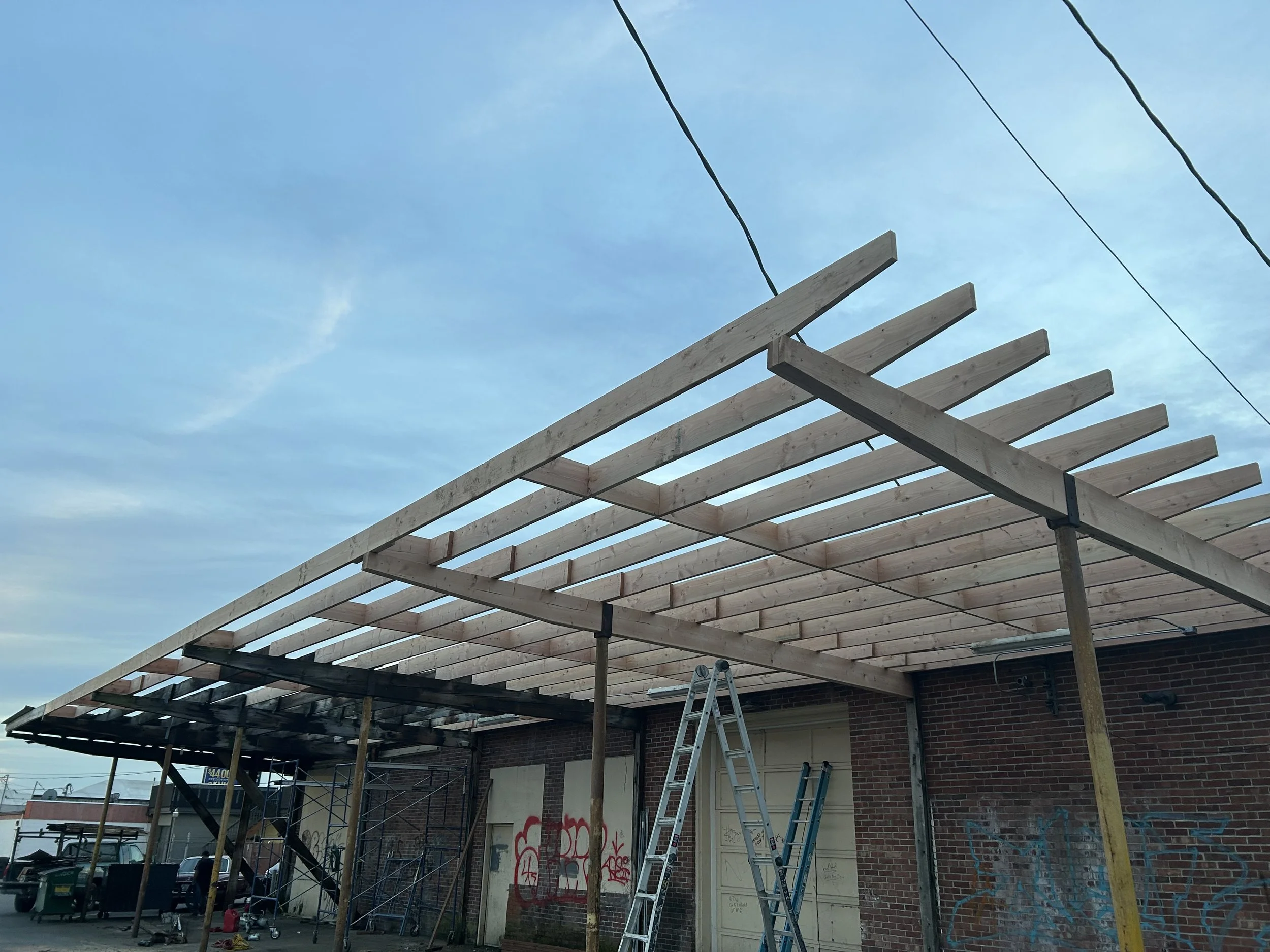 Construction site with wooden beams and scaffolding building a roof or awning outside a brick building with graffiti, under a blue sky.