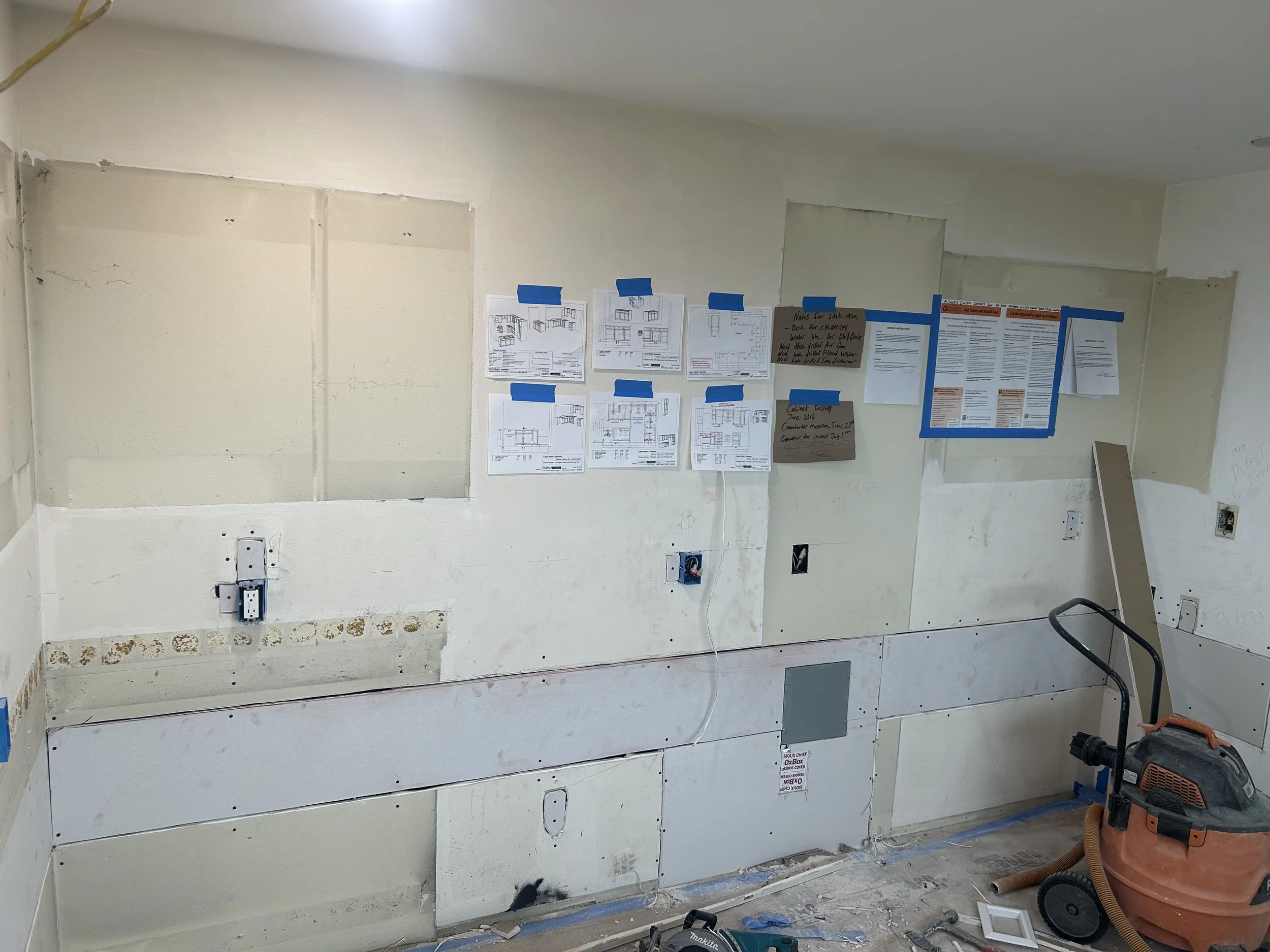 Partially renovated kitchen with exposed drywall, electrical outlets, blue painter's tape, and construction papers on the wall. A shop vacuum is in the corner.