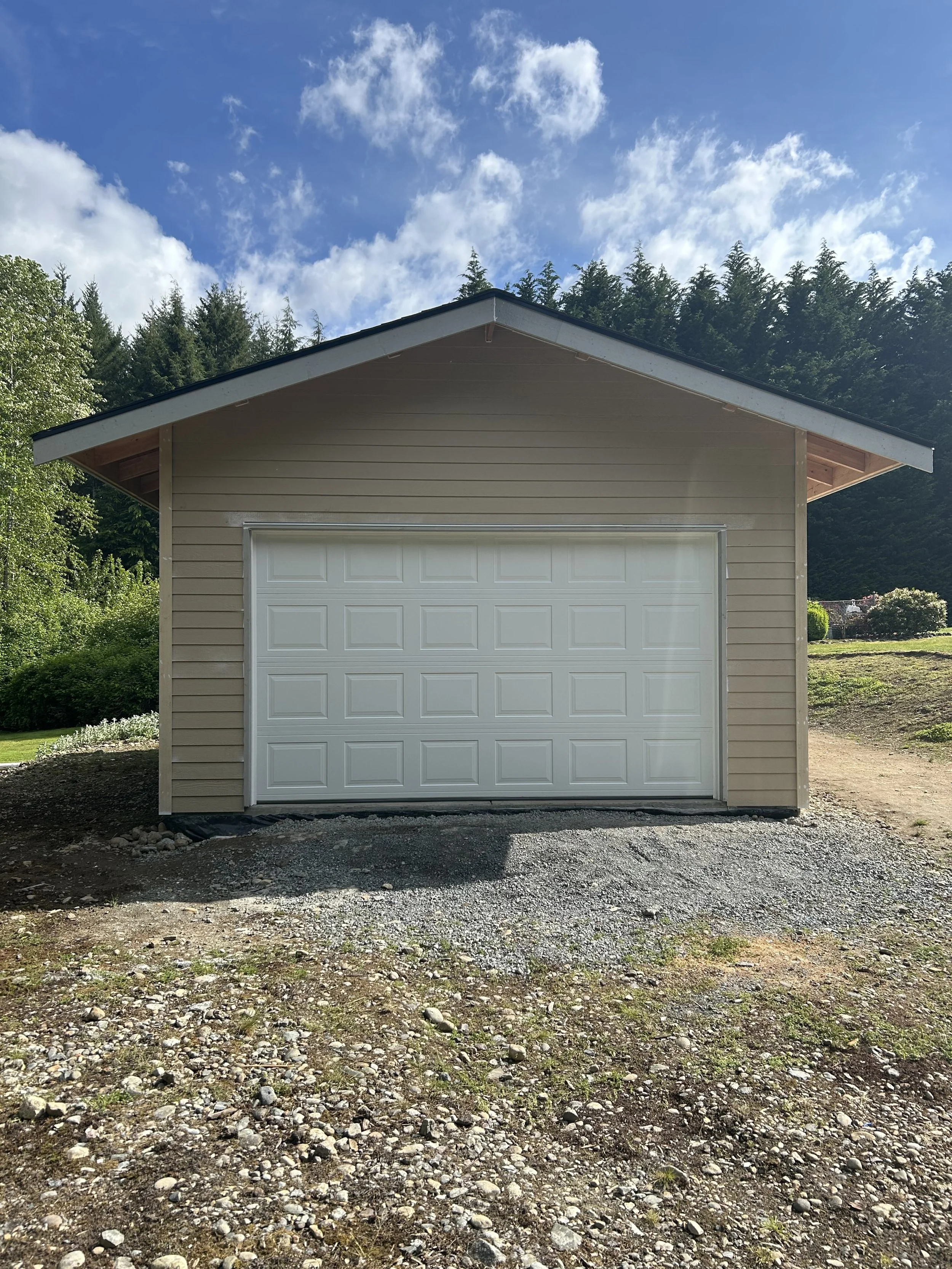 A beige garage with a white door, situated on a gravel area, with a background of green trees and a blue sky with clouds.