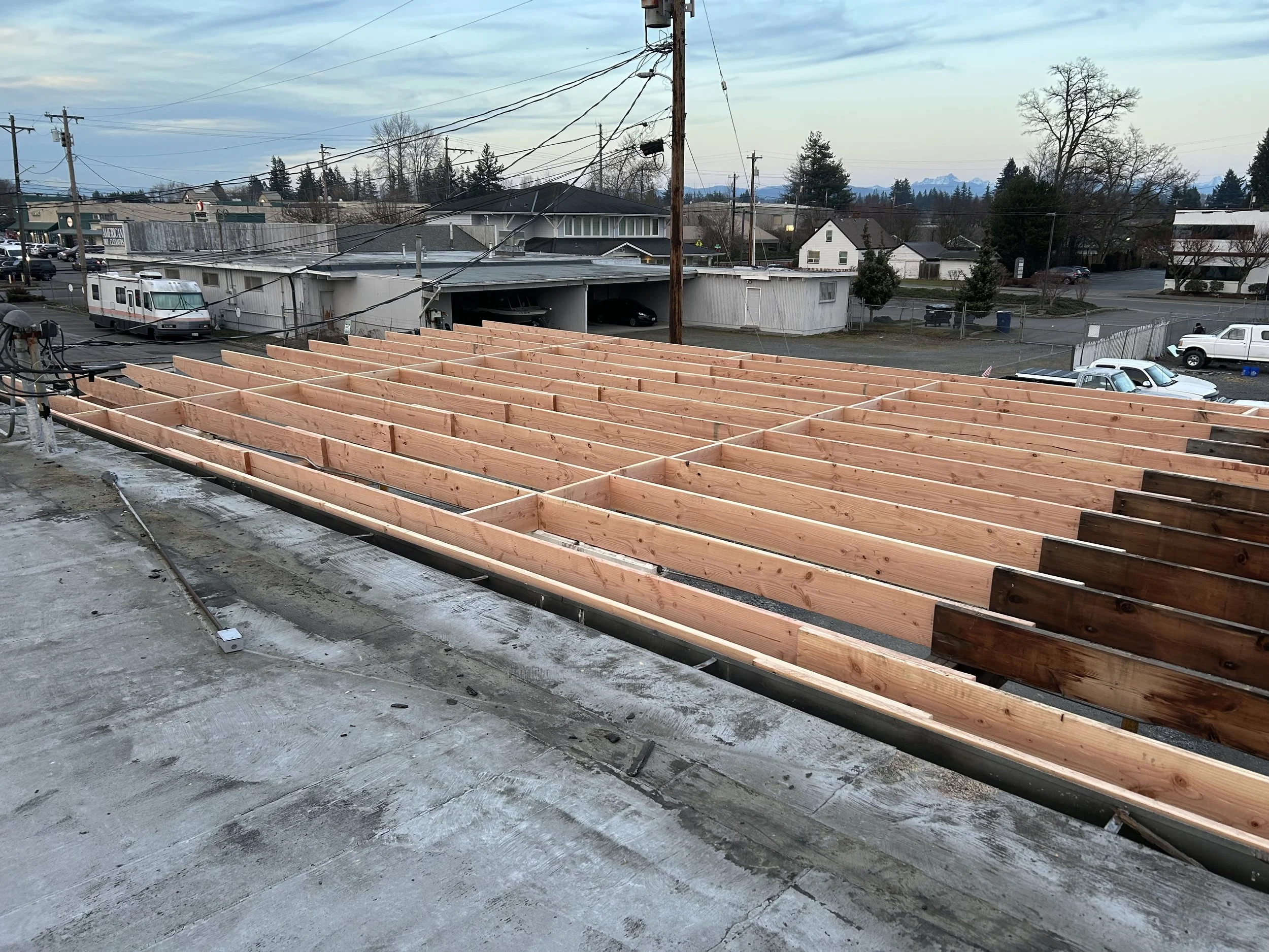 Construction site showing a wooden framework for a building's roof with surrounding urban neighborhood and vehicles in the background.