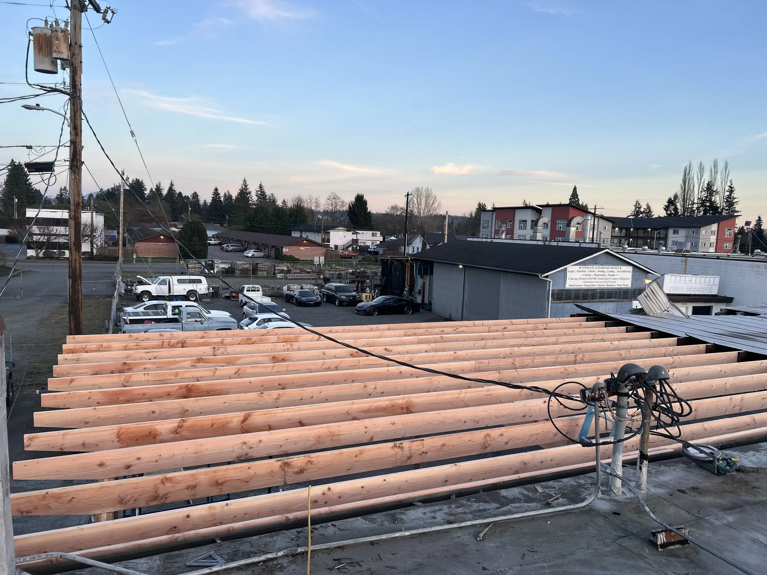 View from a building roof under construction showing wooden beams, power lines, a street with parked cars, and residential and commercial buildings in the background during daytime.
