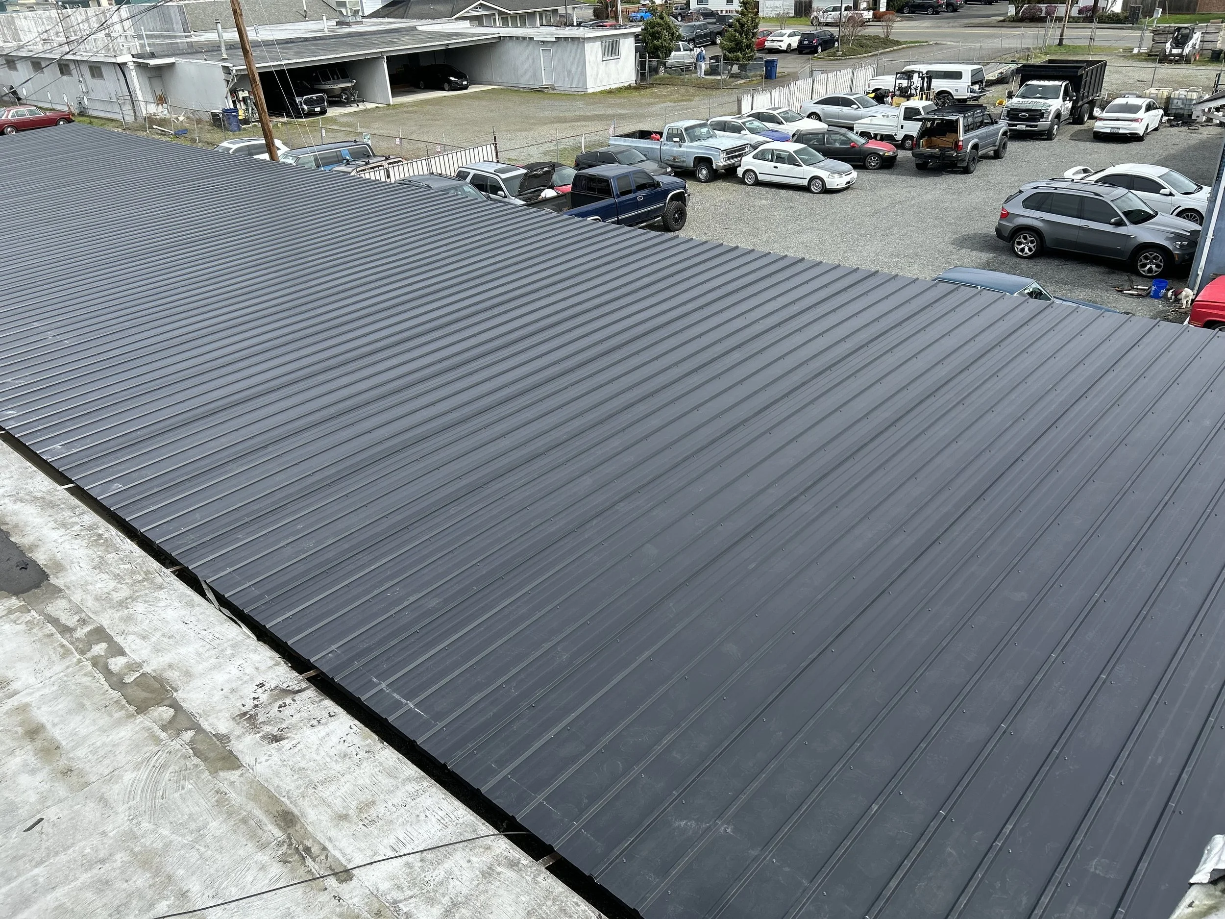 View of a new metal roof installation on a building with a parking lot full of cars and trucks in the background.