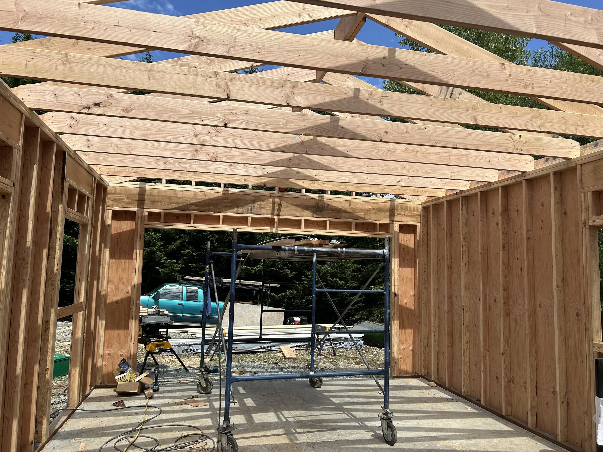 Wooden framing of a building under construction with scaffolding and construction tools inside, and a blue vehicle in the background.